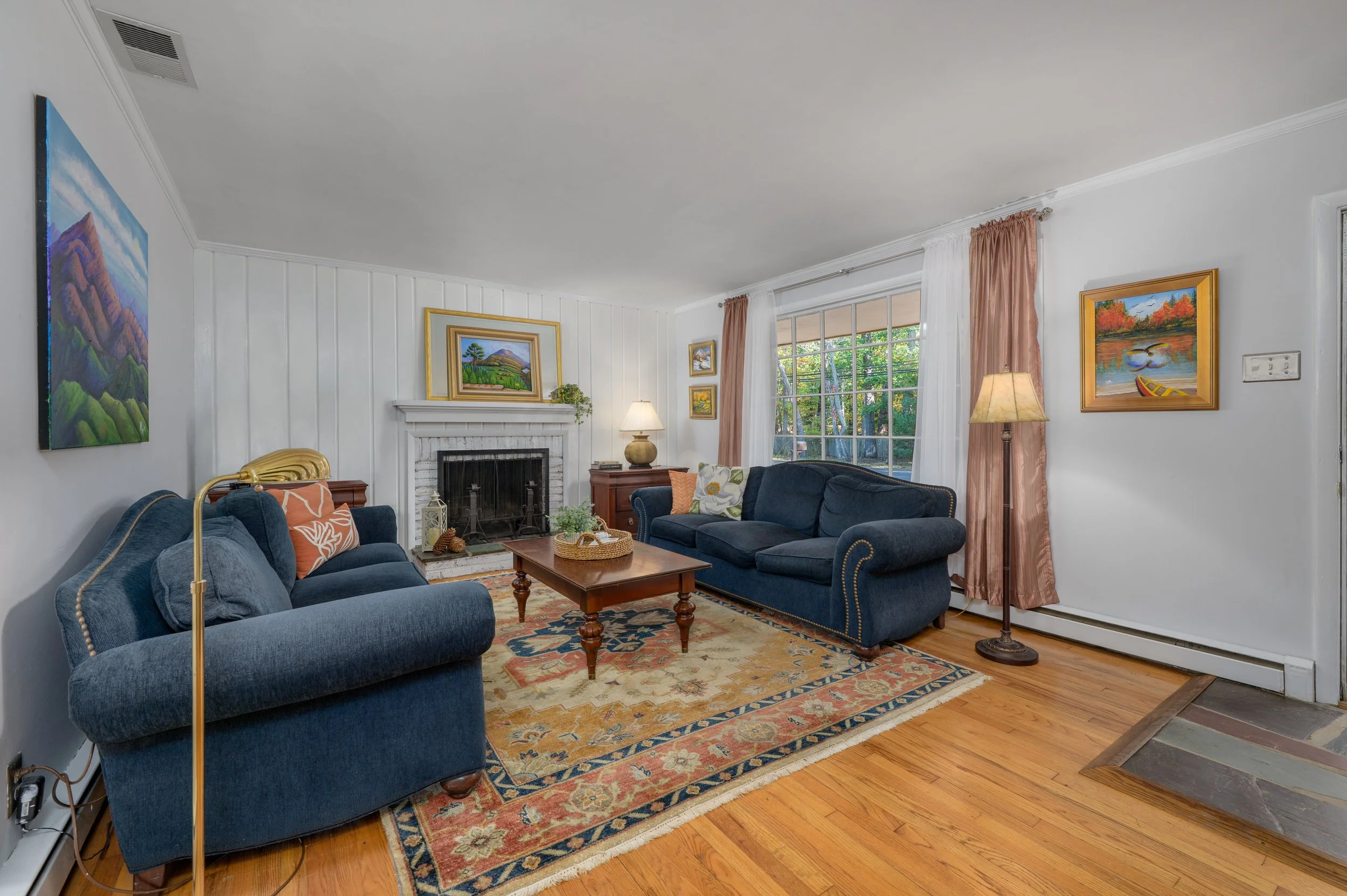 Living room with blue sofas, a wooden coffee table, a fireplace, artwork on white walls, a large window with curtains, and hardwood floors.