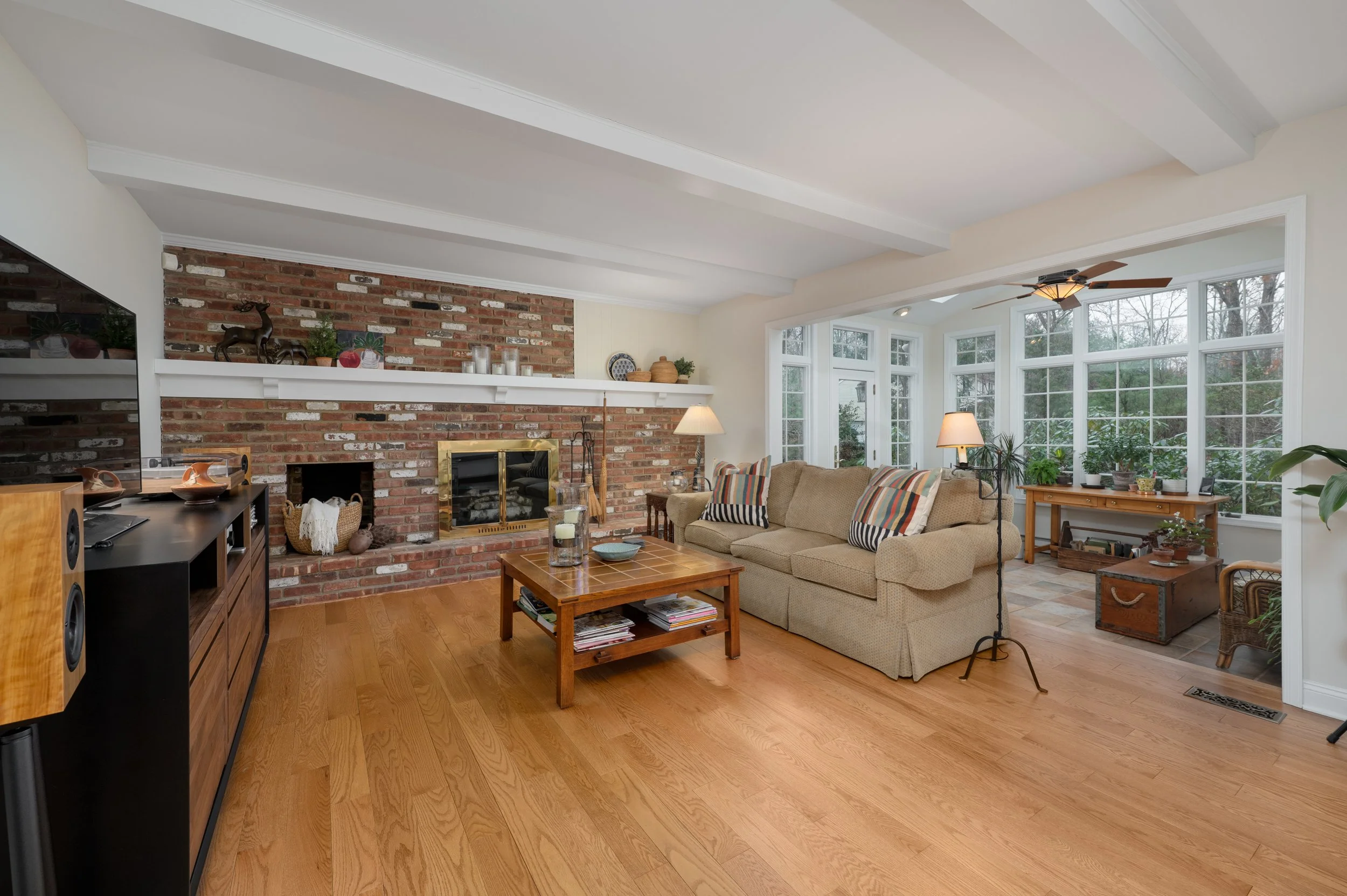 Living room with brick fireplace, beige sofa with striped pillows, wooden coffee table, large bay window with potted plants, hardwood floors, and ceiling fan.