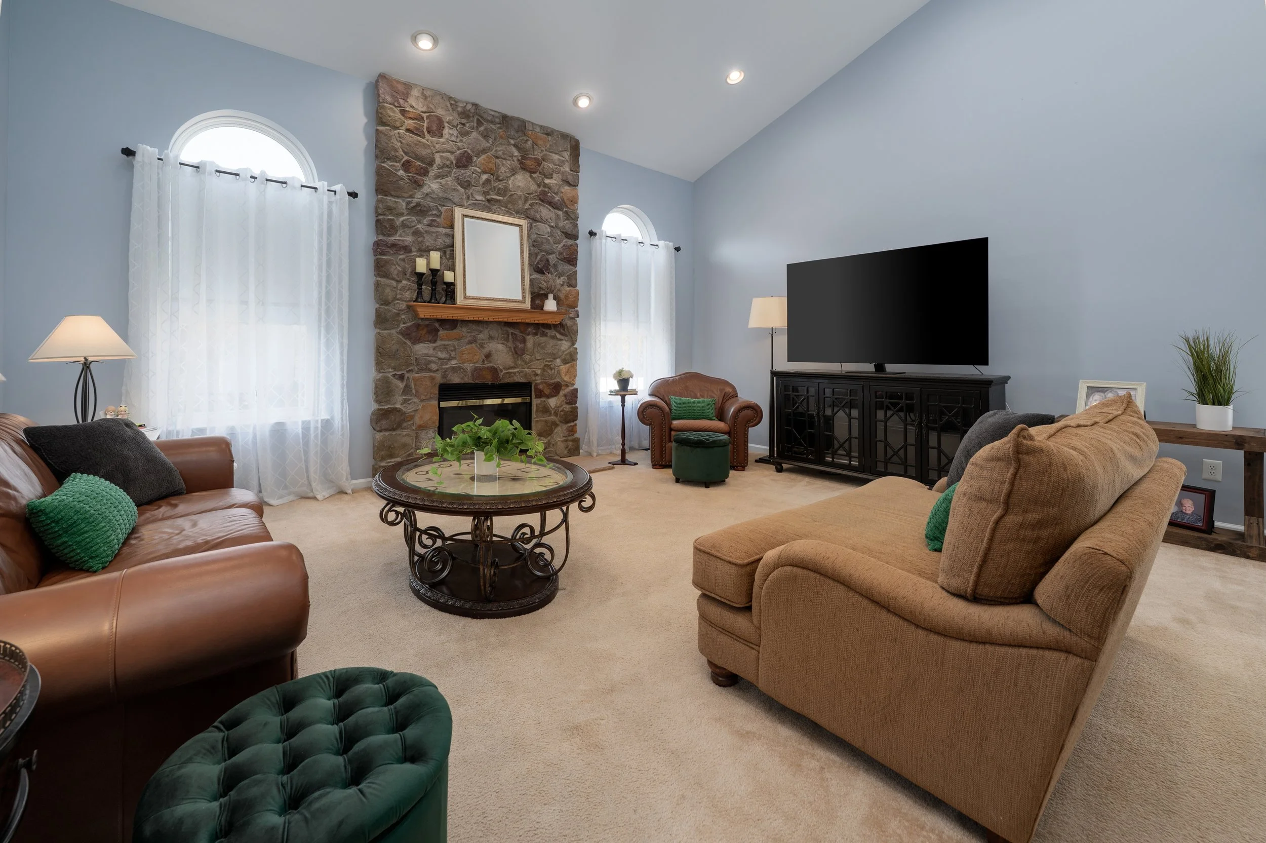 Living room with beige carpet, tan and brown sofas, a stone fireplace, a black TV on a cabinet, and decorative pillows, with two windows with white curtains and a small table with a plant.