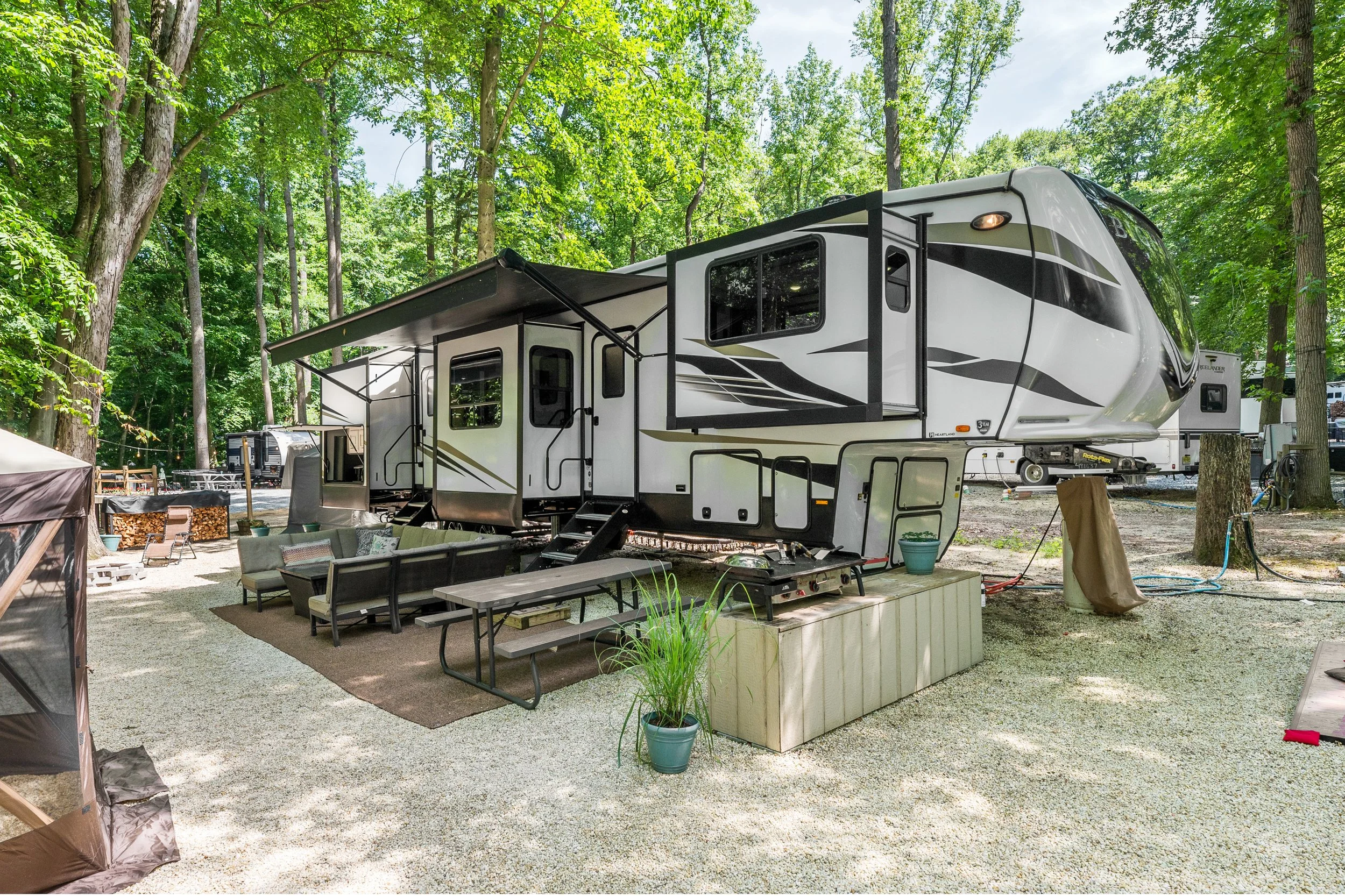 A modern recreational vehicle (RV) parked in a wooded campsite with outdoor furniture and potted plants.