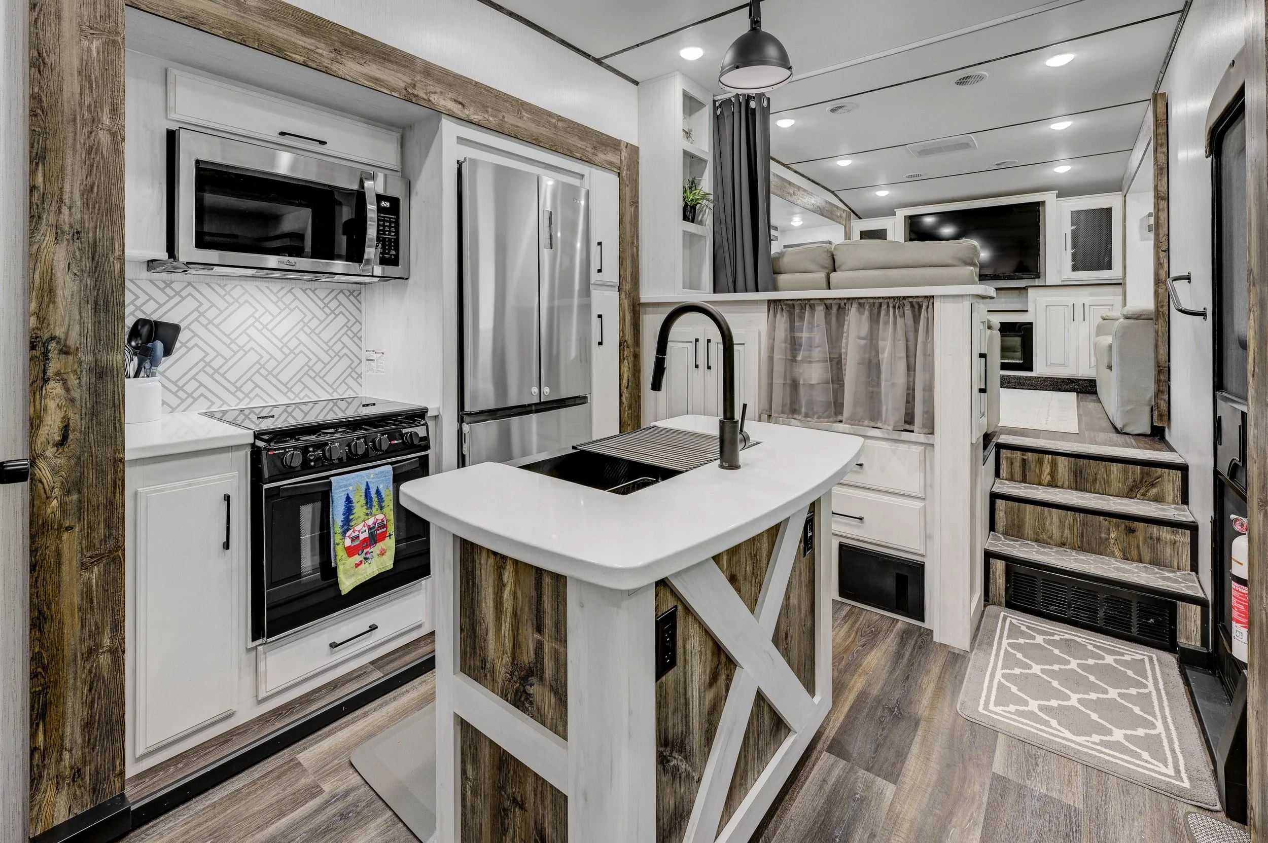 Compact kitchen with black stove, microwave, stainless steel refrigerator, white cabinetry with black handles, and a small kitchen island with a black faucet. Visible living space with stairs, TV, white seating, and wood accents.