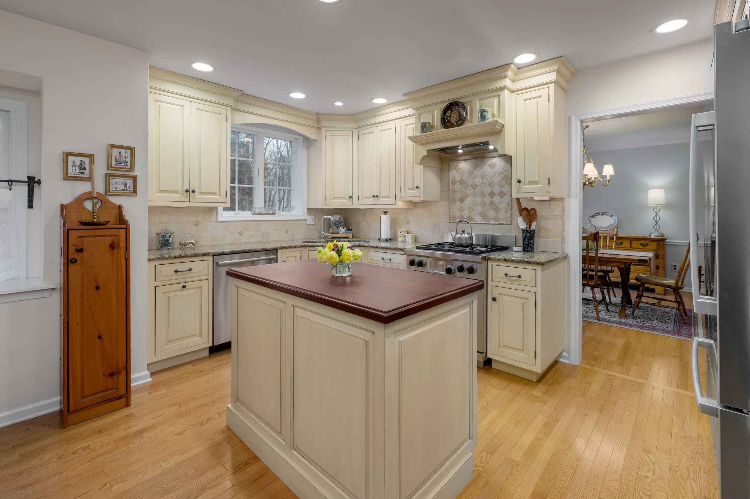 Kitchen with cream-colored cabinets, a wooden kitchen island with a dark wood top, and hardwood floors. There is a window above the sink, a stove with a tiled backsplash, and a small dining area visible through an open doorway.