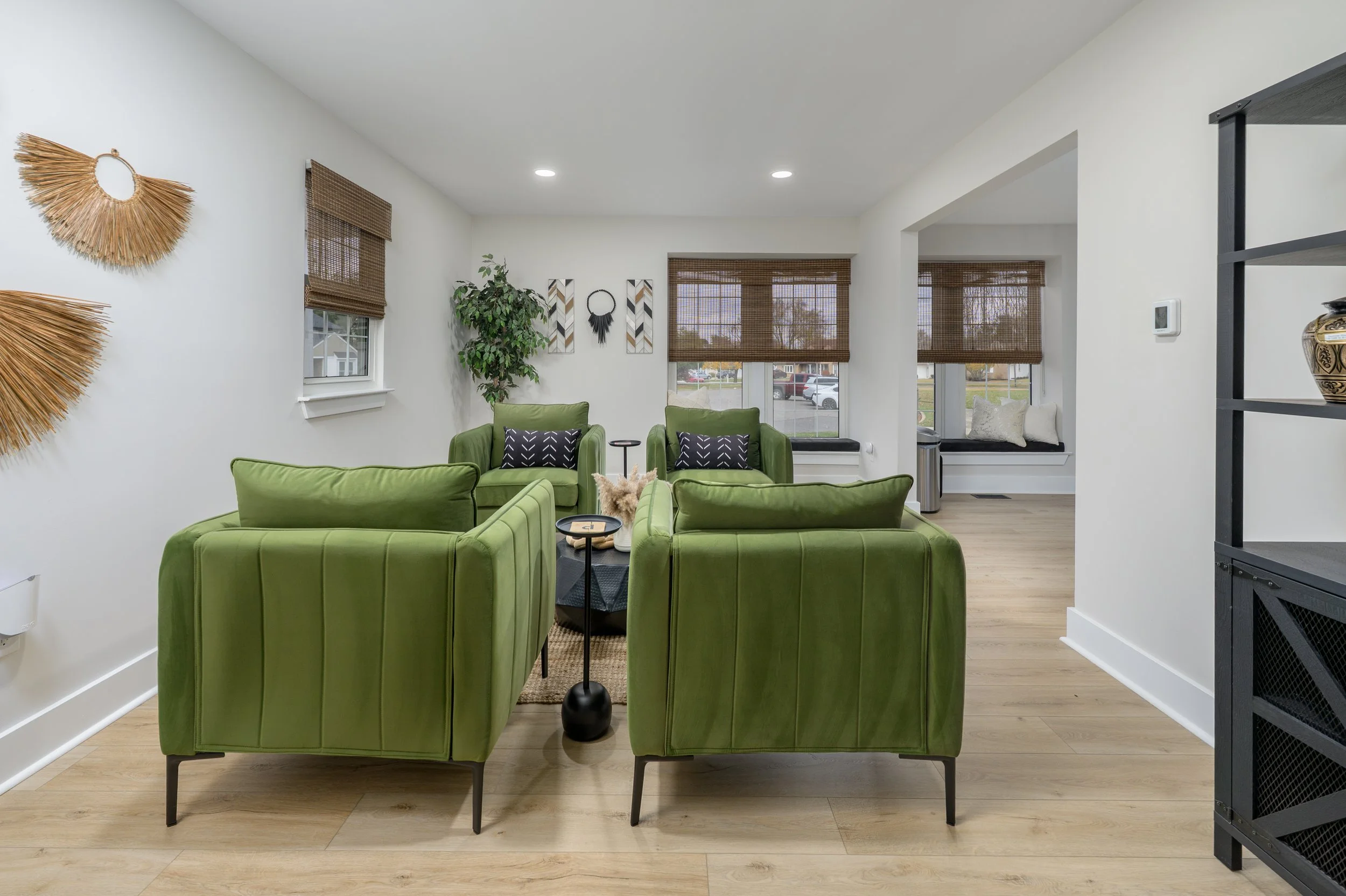 Living room with four green armchairs arranged around a coffee table, decorative wall art, and large windows with brown woven blinds.