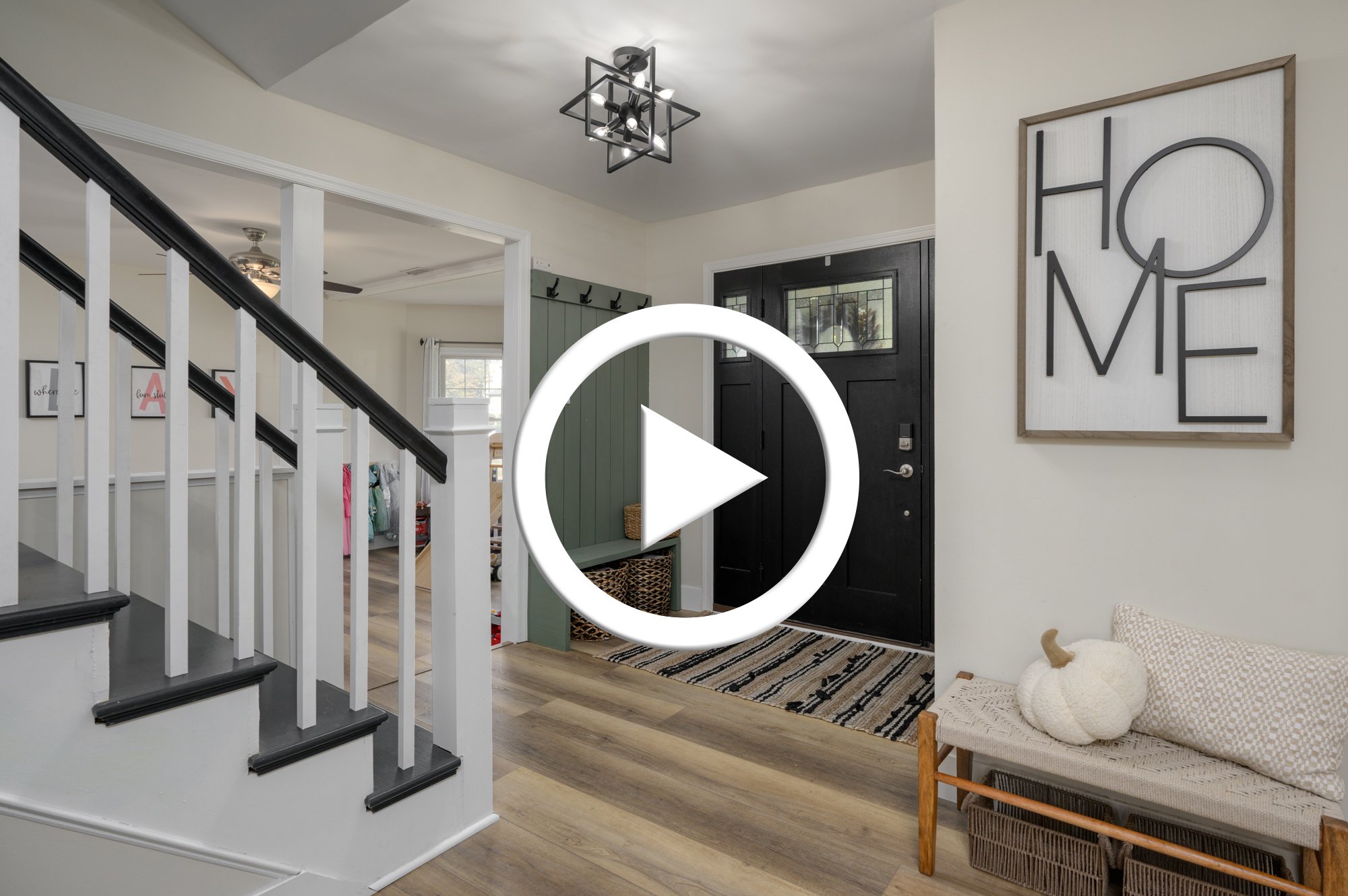 Entrance area of a home with a black front door, a wall sign saying 'HOME', a bench with a decorative pumpkin, a striped rug, and a foyer with hooks and green wall paneling.