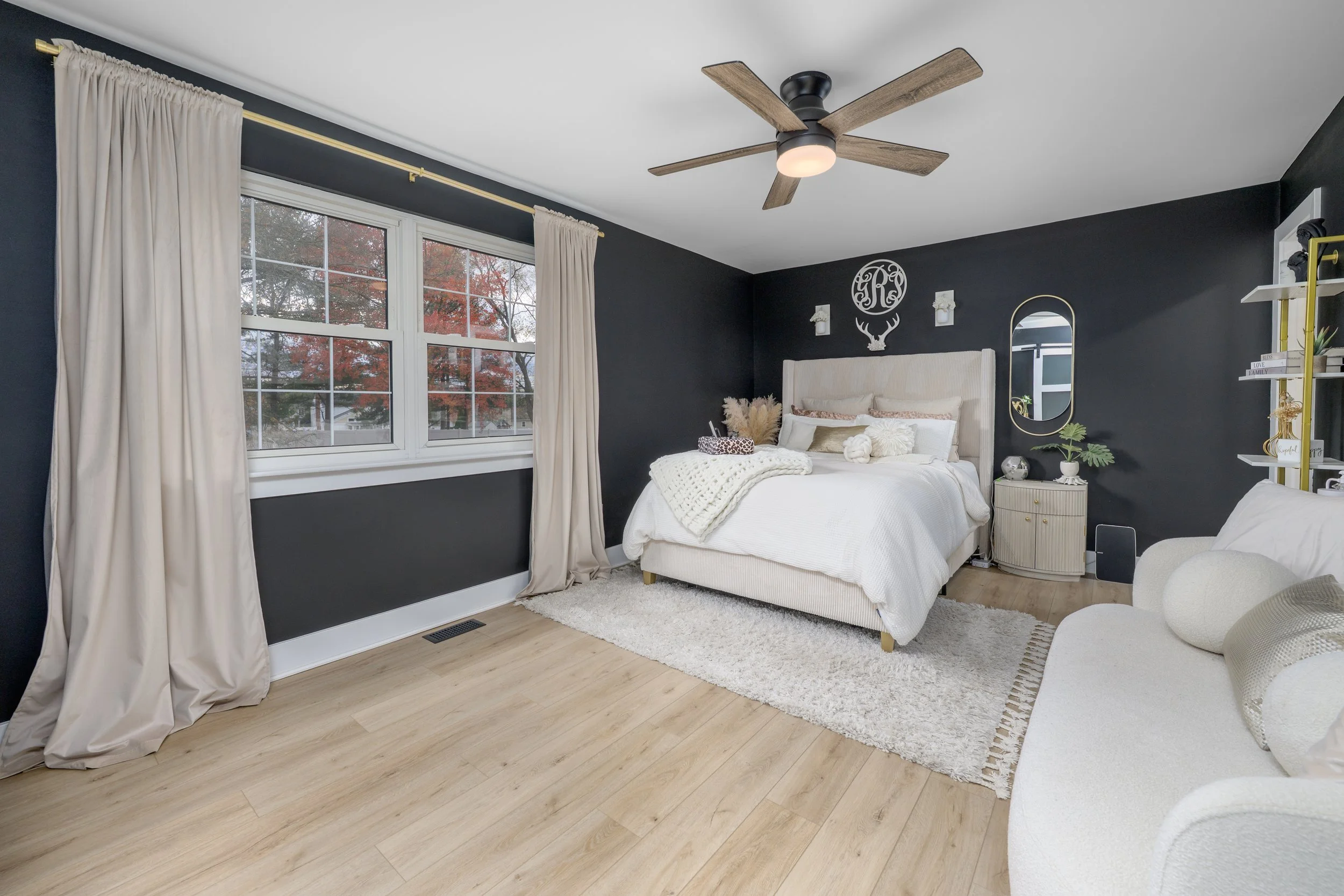 A bedroom with black walls, a large window with cream-colored curtains, a ceiling fan, a white bed with decorative pillows, a small side table with plants and decorations, a white sofa, and a rug.
