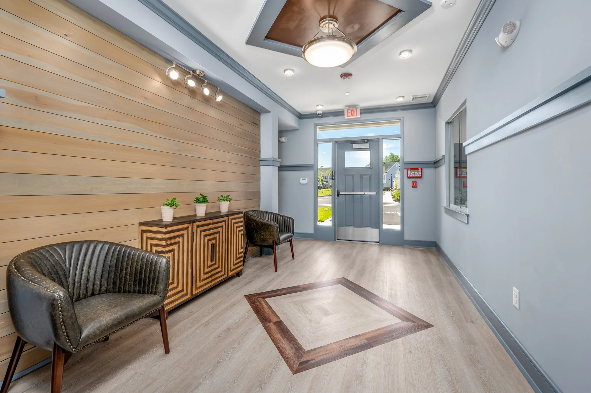 Interior of a modern building lobby with gray walls, a wooden accent wall, two black chairs, a wooden cabinet, green plants, and glass doors leading outside.