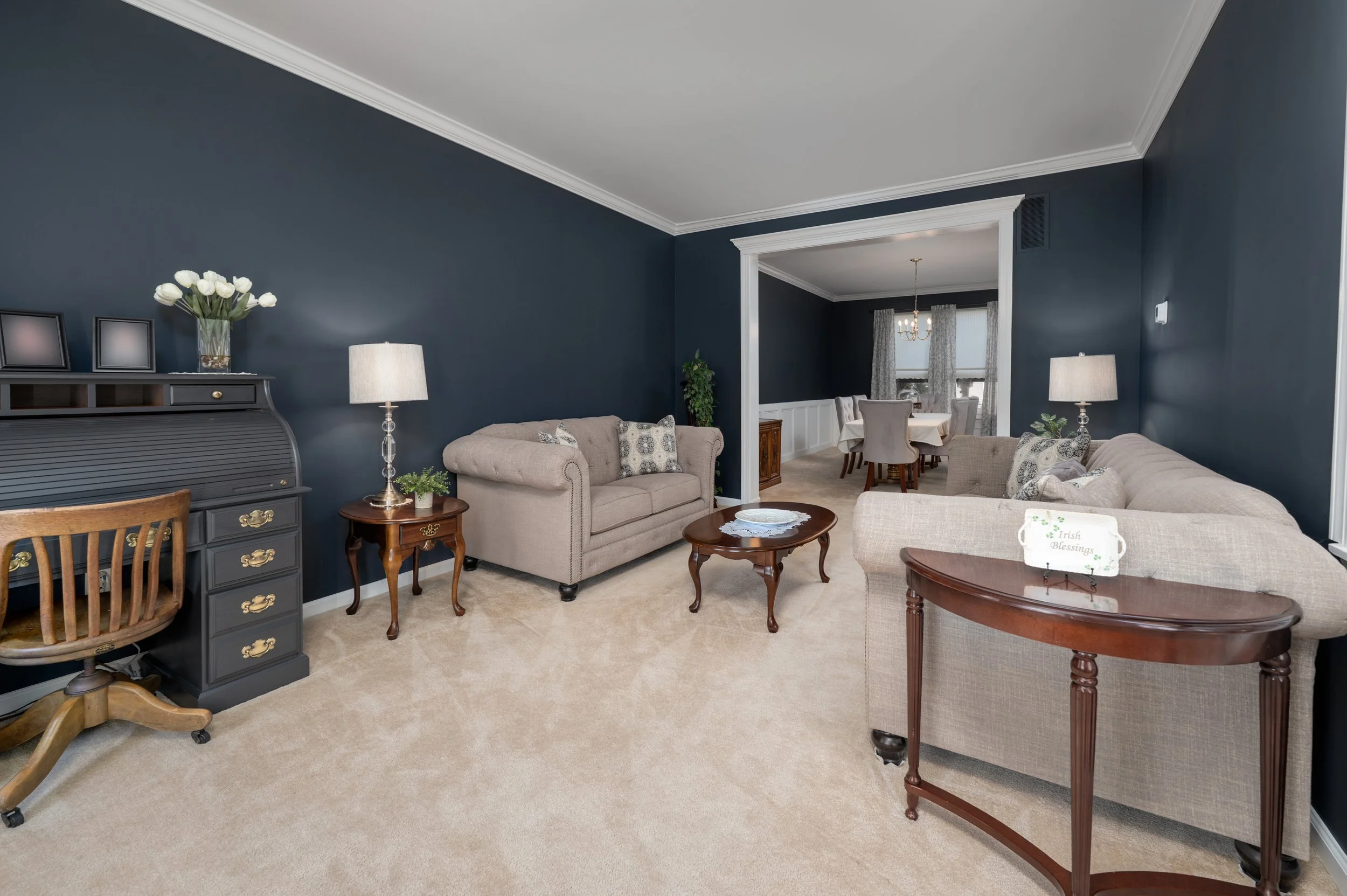 Living room with beige and gray sofas, dark blue walls, beige carpet, wooden side tables, lamps, and a view into the dining area with a chandelier and window curtains.