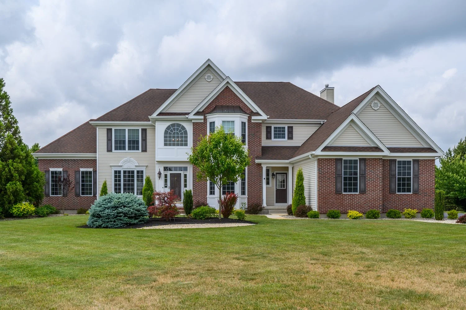 A large two-story house with brick and beige siding exterior, multiple windows, and a landscaped front yard with grass, bushes, and small trees.