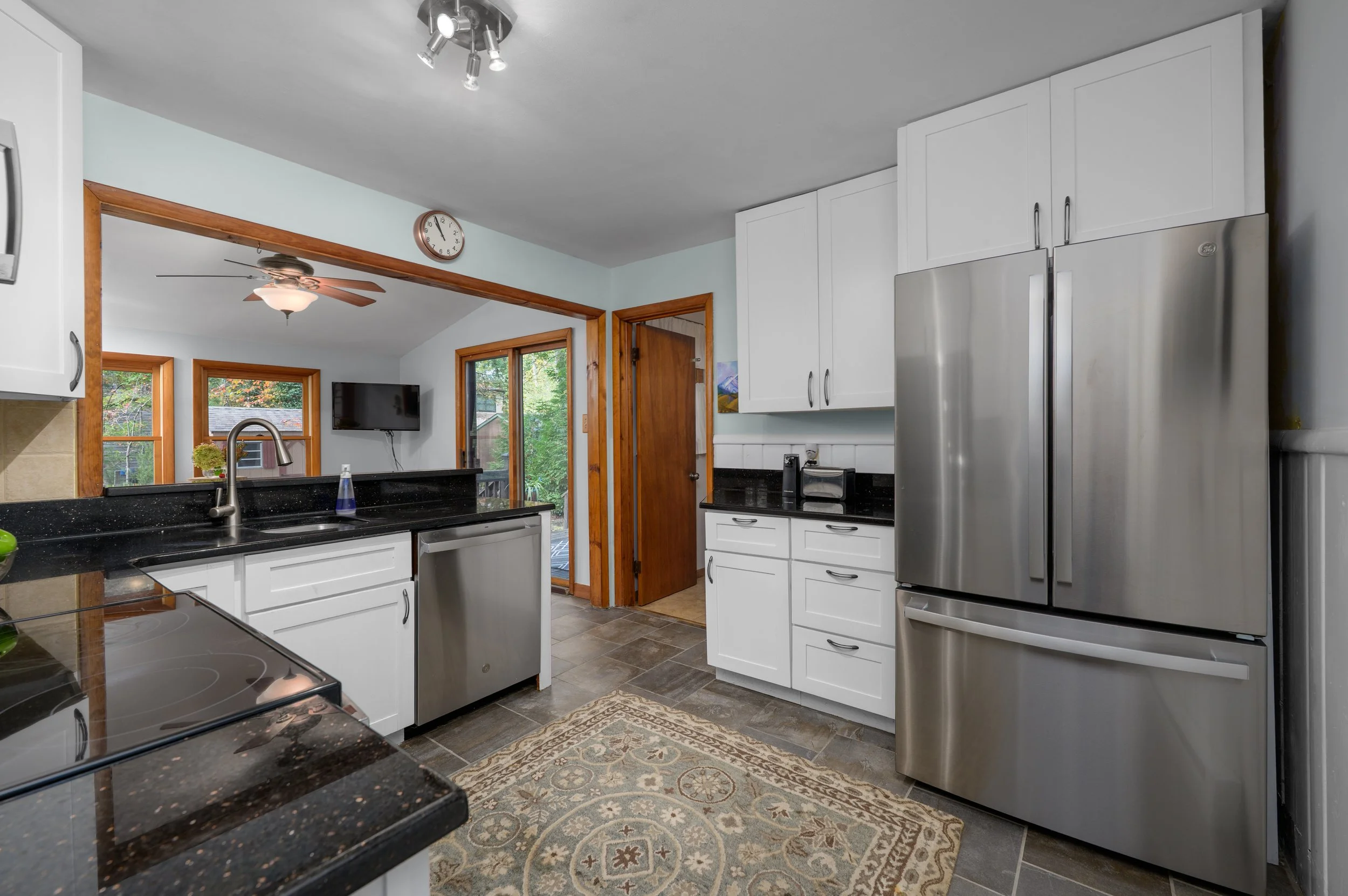 Kitchen with white cabinets, black countertops, stainless steel refrigerator, and a patterned rug on the tiled floor. Visible windows and a door leading outside, with a view of greenery.