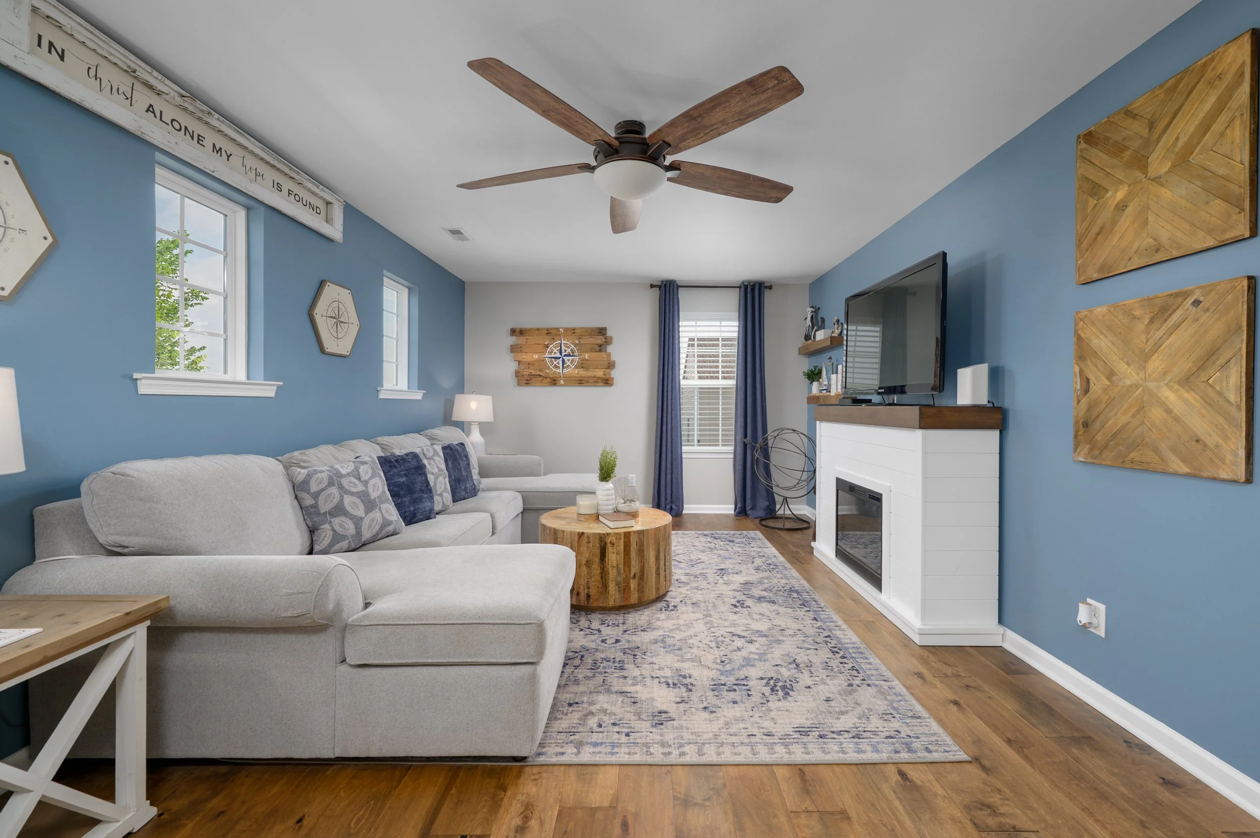 Living room with a blue accent wall, white sofa, wooden coffee table, and a fireplace with a TV mounted above. Decorations include wall art, curtains, and a patterned rug.