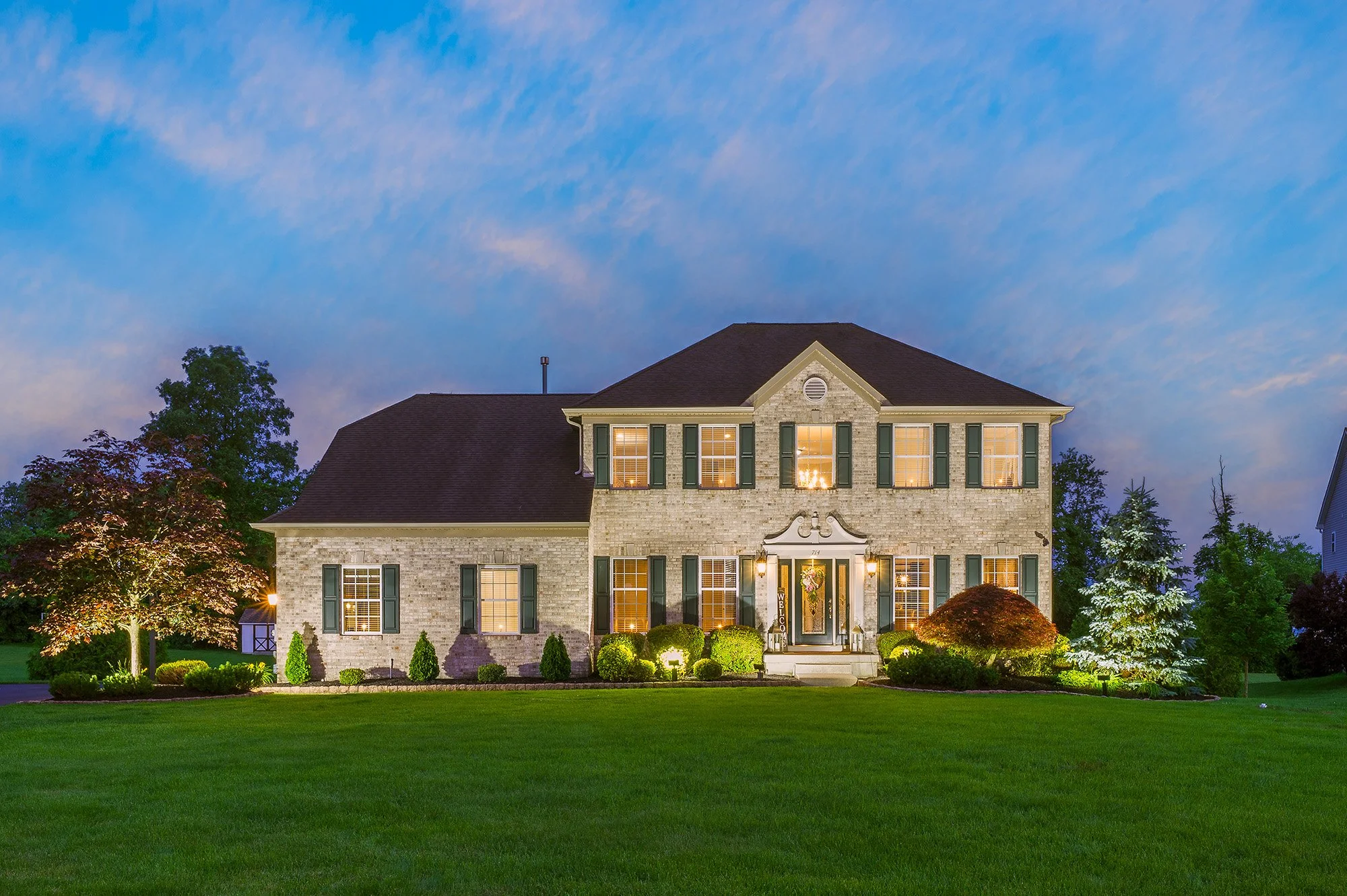 A well-lit two-story house with a brick exterior, green shutters, and a dark roof, illuminated at dusk, surrounded by a lush green lawn and landscaped bushes and trees.