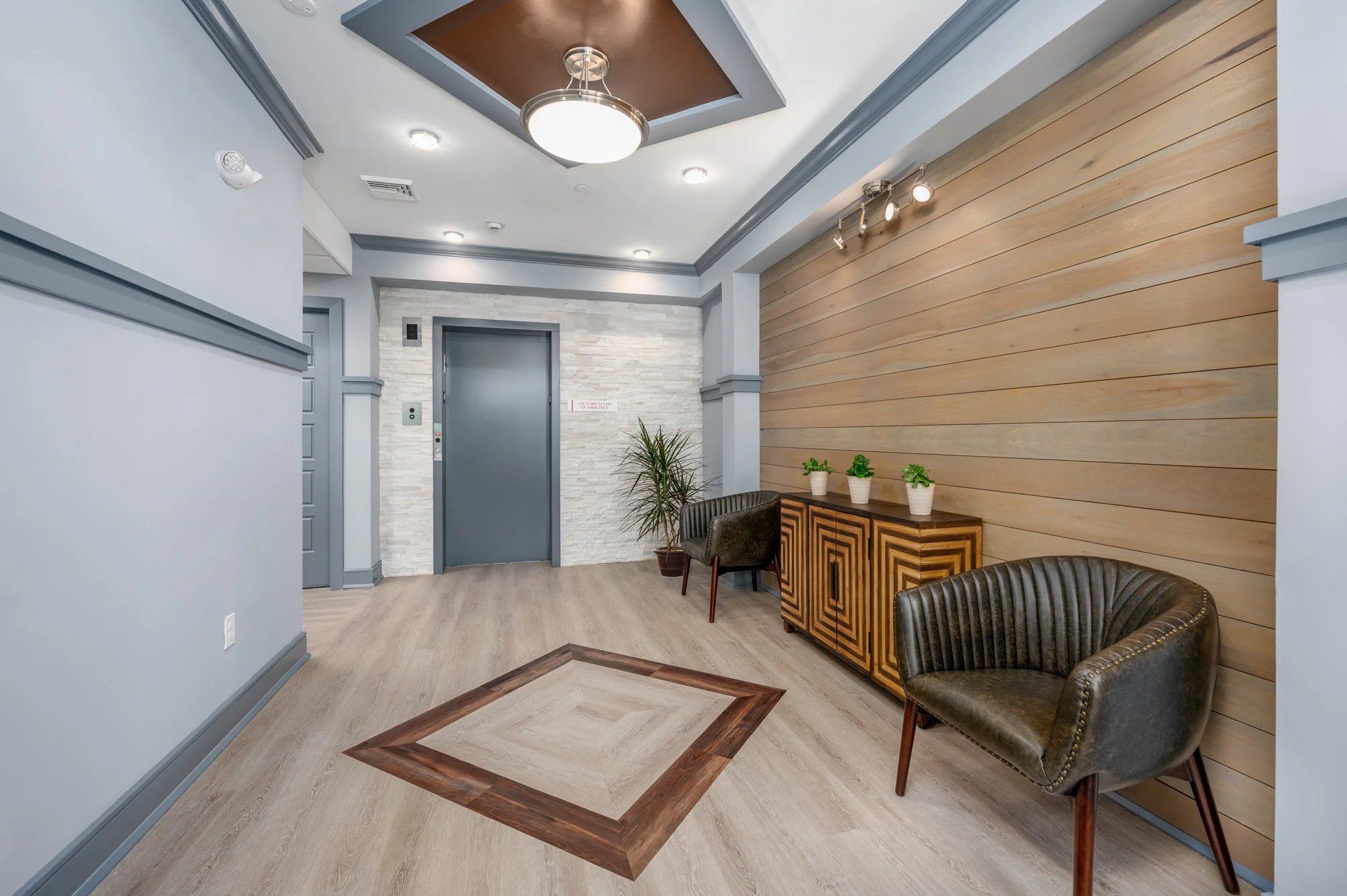 Modern lobby with grey elevators, wooden accent wall, two black leather chairs, a wooden cabinet with three potted plants, wood flooring, and ceiling lighting.