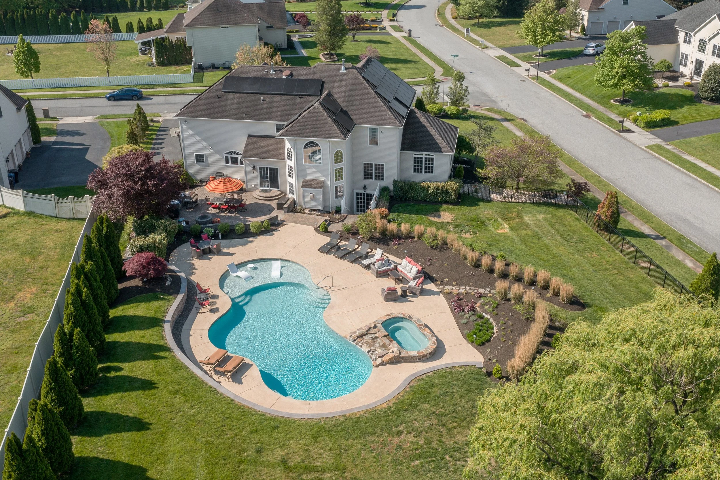 Aerial view of a backyard with a swimming pool, hot tub, and outdoor seating area, adjacent to a white house with solar panels on the roof, surrounded by a landscaped garden and fenced yard in a suburban neighborhood.