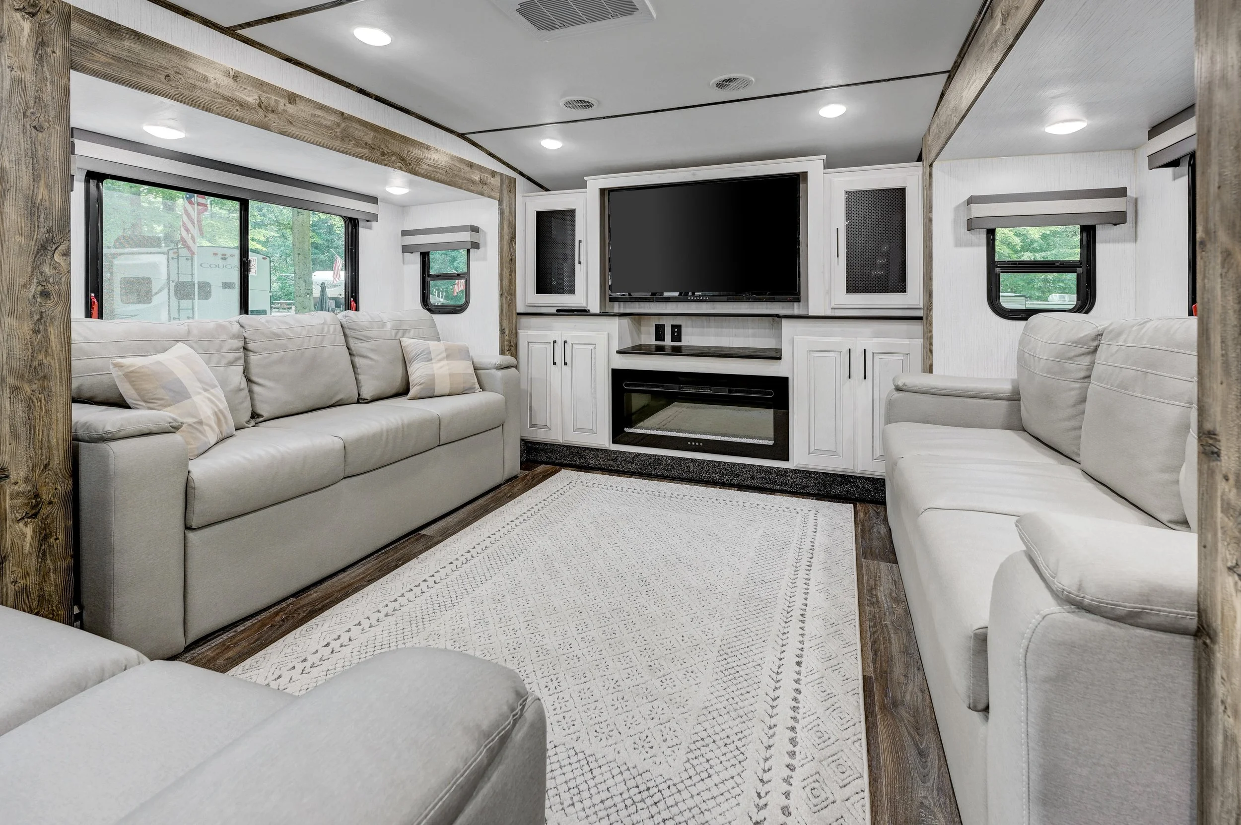Living room with two beige sofas, a white built-in entertainment center with a large flat-screen TV, wood trim accents, windows with blinds, and a white patterned area rug.