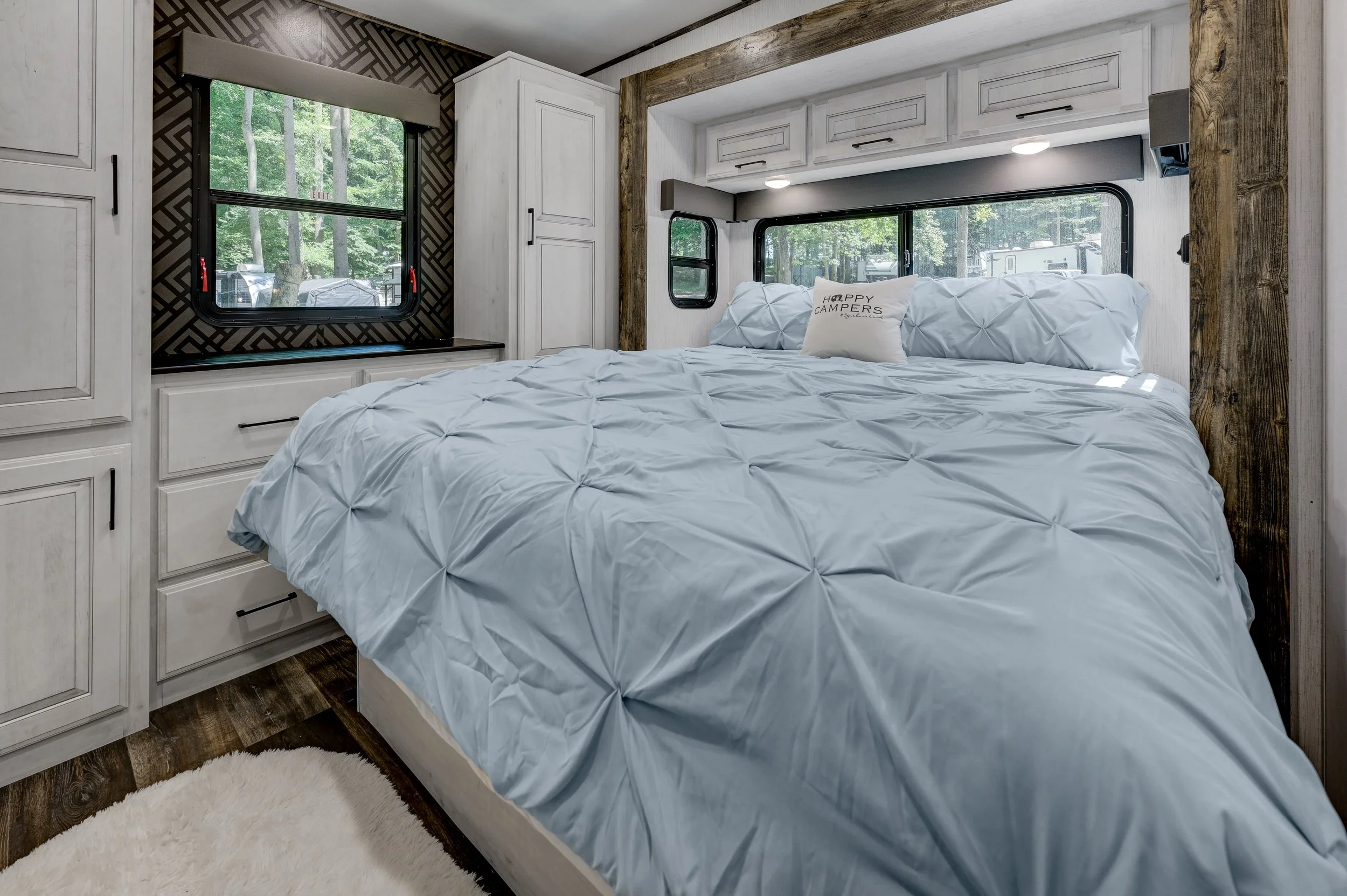 Interior of an RV bedroom with a large bed featuring white comforter and pillows, surrounded by white cabinetry, windows, and wooden accents, with a view of trees outside.