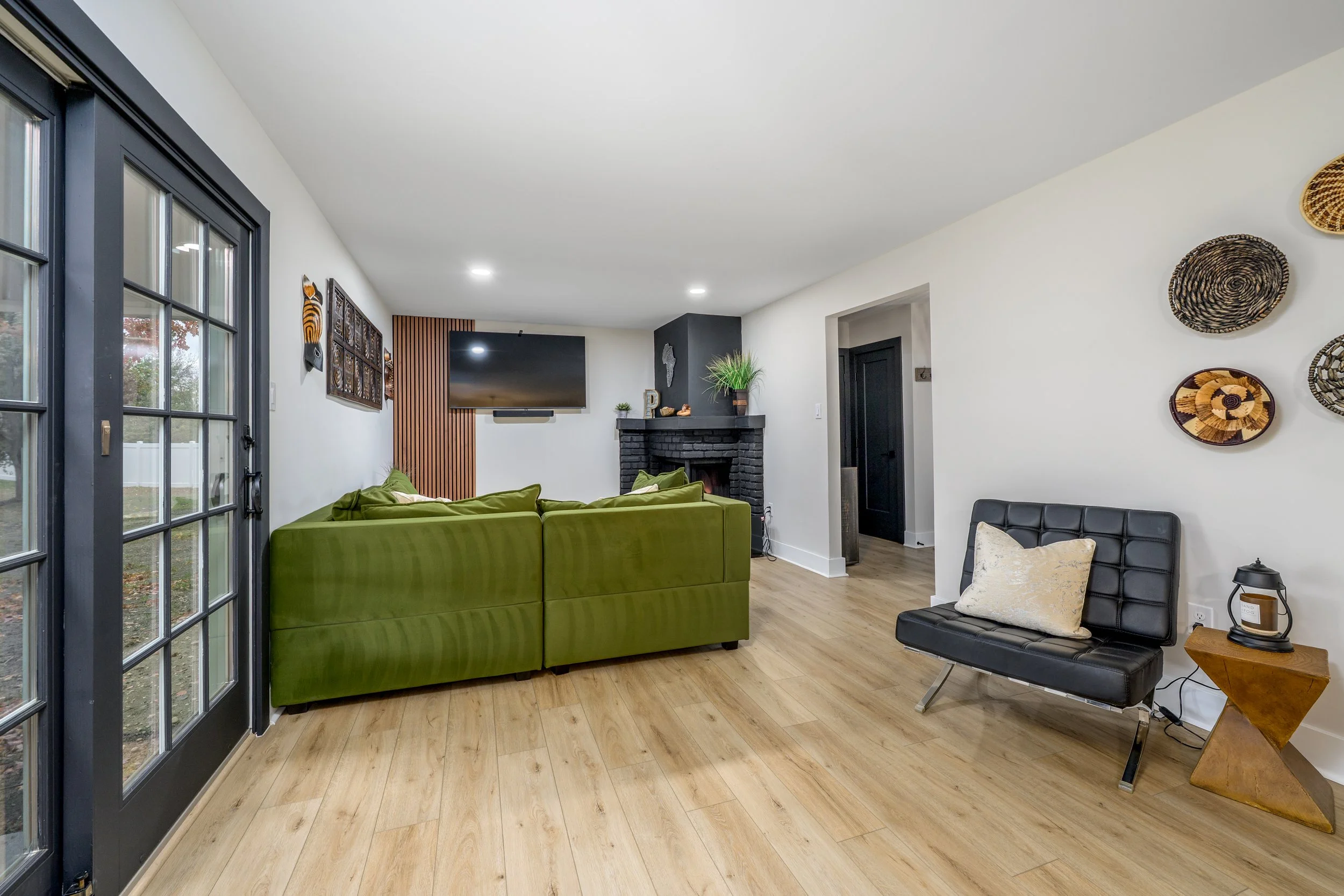 Living room with black-framed glass doors, green sofa, black chair with pillow, wall decorations, mounted TV, fireplace, and hardwood floors.
