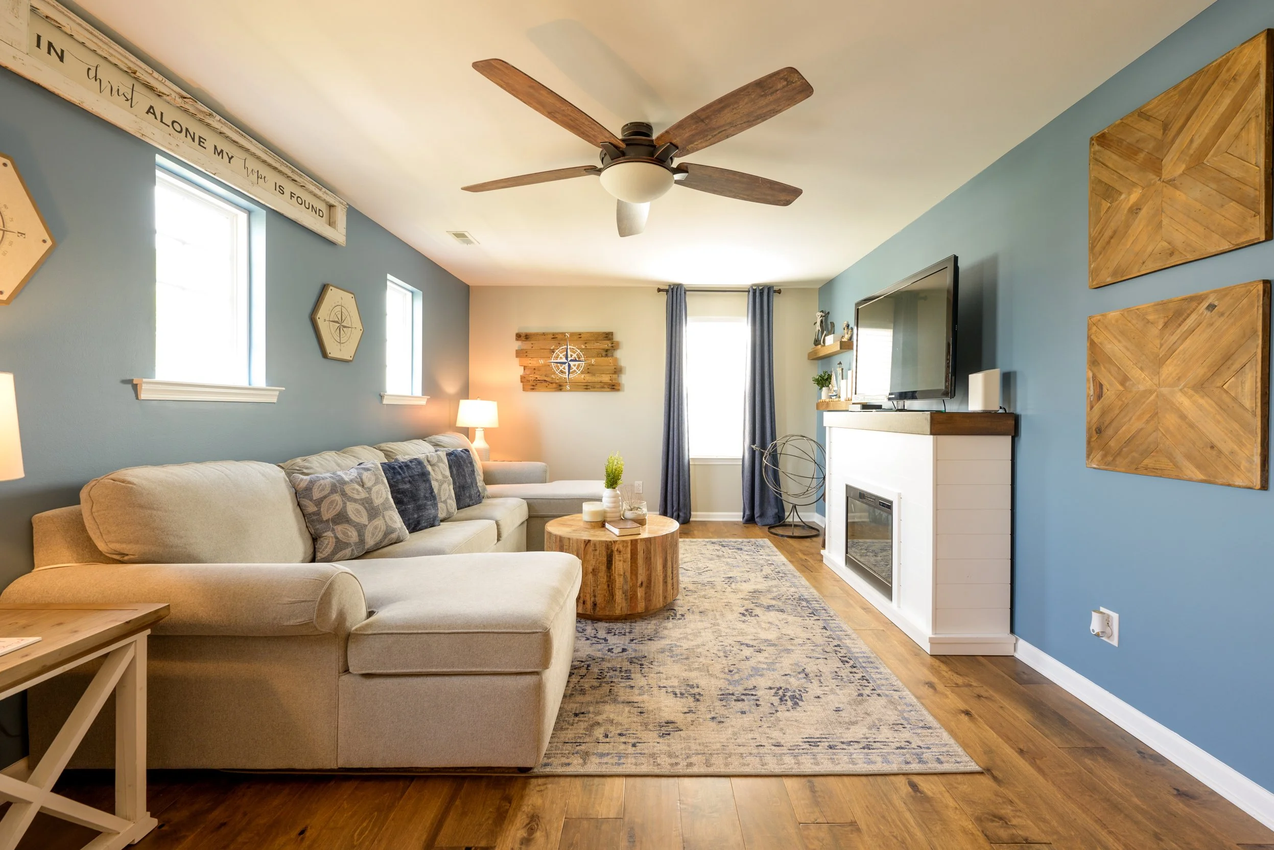 Living room with beige sectional sofa, wooden coffee table, blue accent wall, wall art, and a ceiling fan.