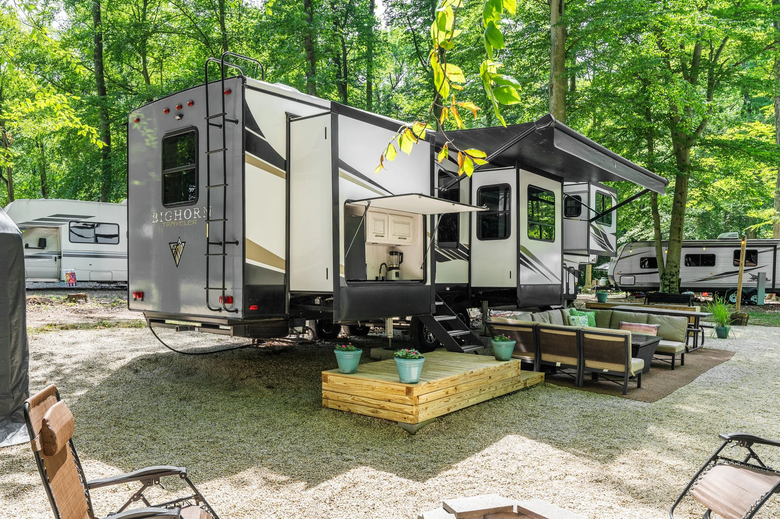 A modern travel trailer with an outdoor seating area on a gravel campsite surrounded by green trees.