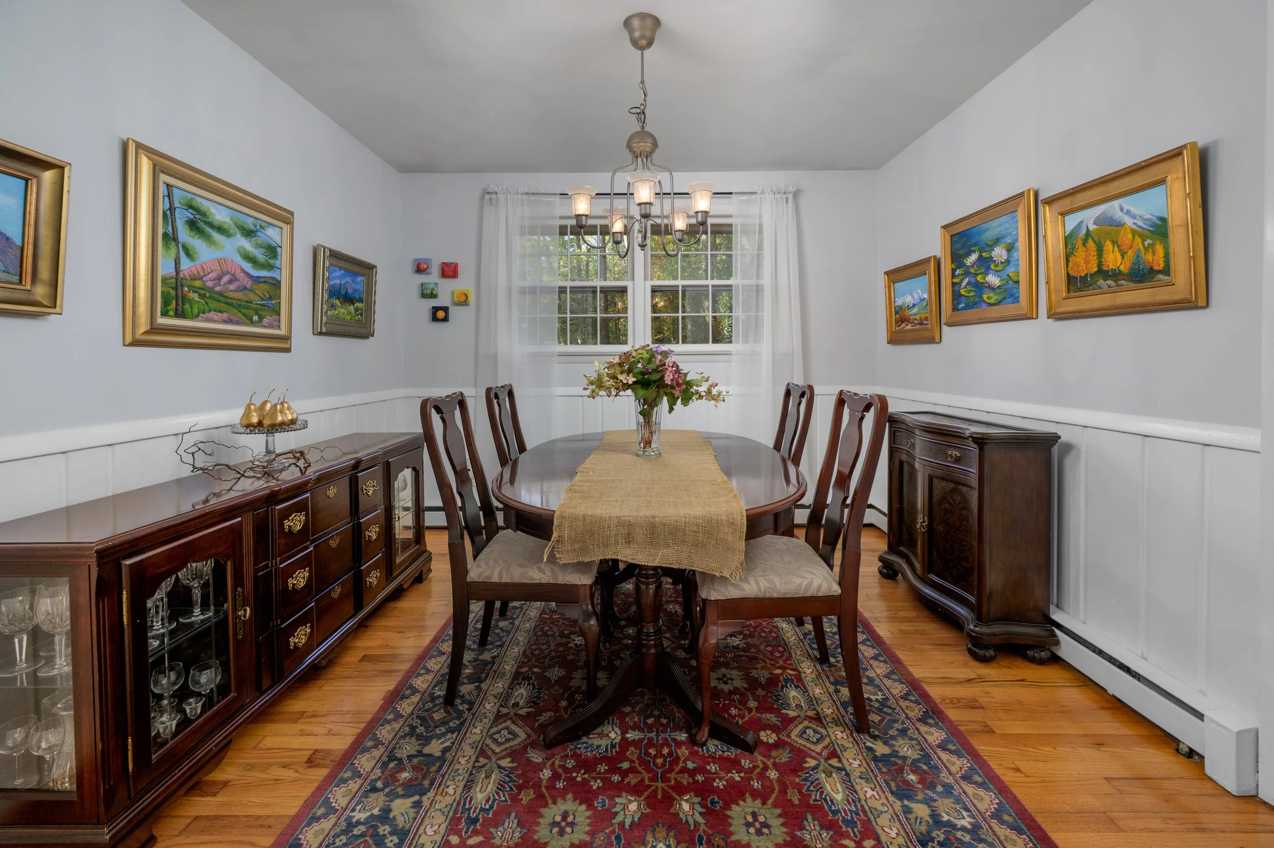 Dining room with a wooden table and six chairs, a chandelier, framed landscape paintings on white walls, a window with white curtains, a dark wood sideboard, and a floral centerpiece, with a colorful rug on hardwood floor.