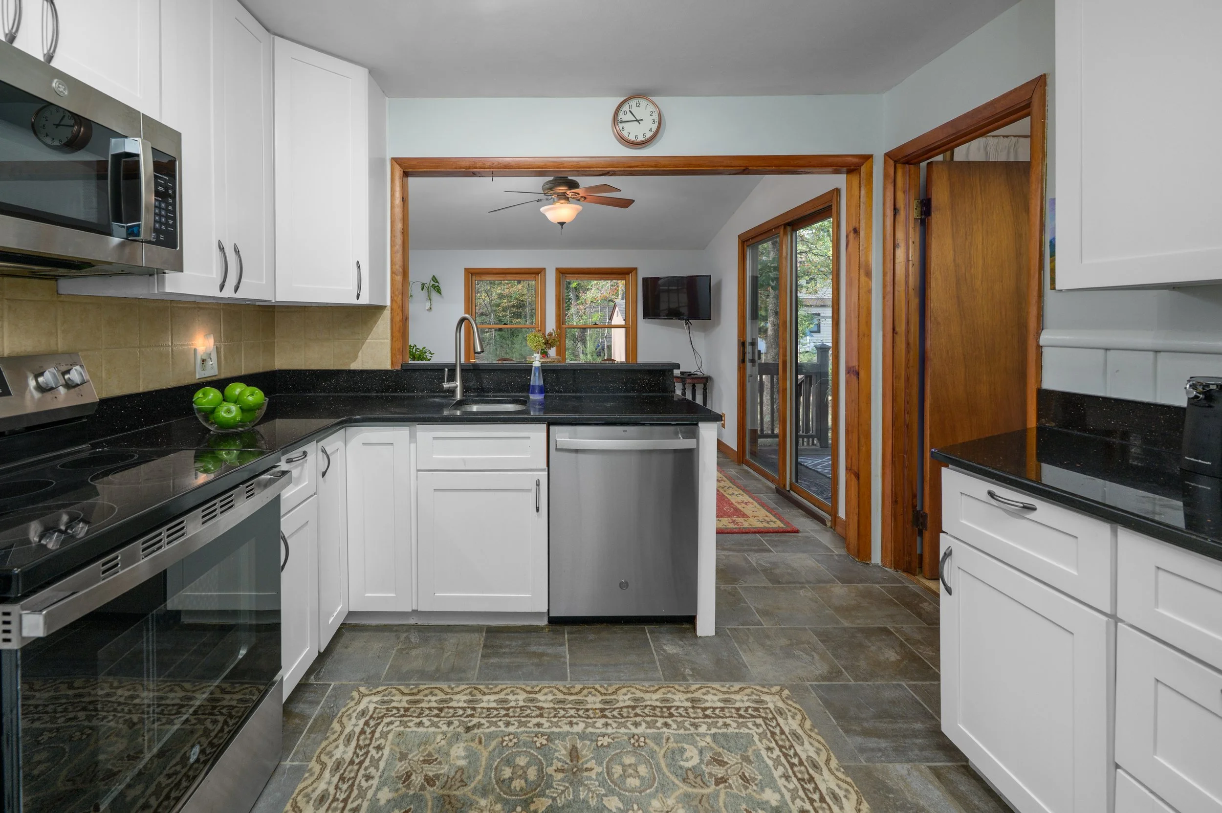 Kitchen with black countertops, white cabinets, stainless steel appliances, a bowl of green apples, beige backsplash, and a view into a living room with a ceiling fan, windows, a wall-mounted TV, and a sliding glass door leading outside.