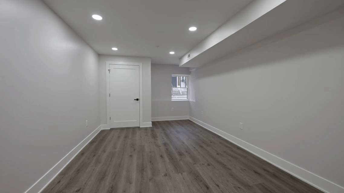Empty room with white walls, hardwood flooring, a white door, a small window, and ceiling lights.