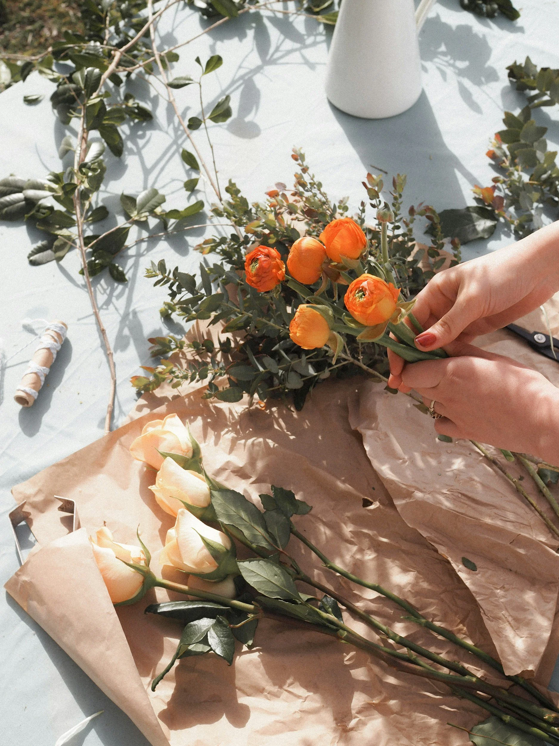 Person arranging orange and cream-colored roses with greenery on a table, surrounded by floral supplies and wrapping paper.