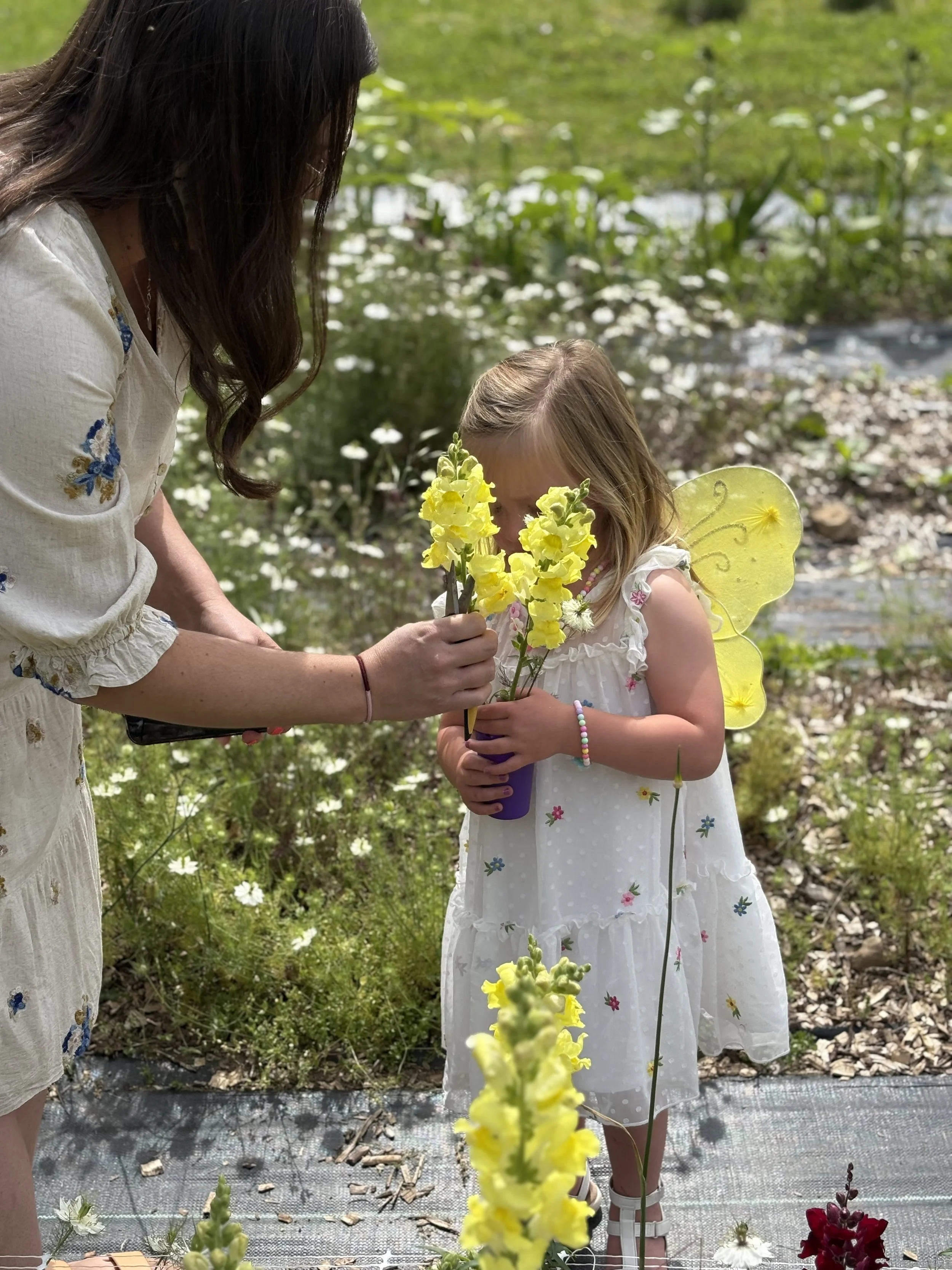A woman helps a young girl with fairy wings hold a purple flower pot filled with yellow flowers outdoors in a garden.