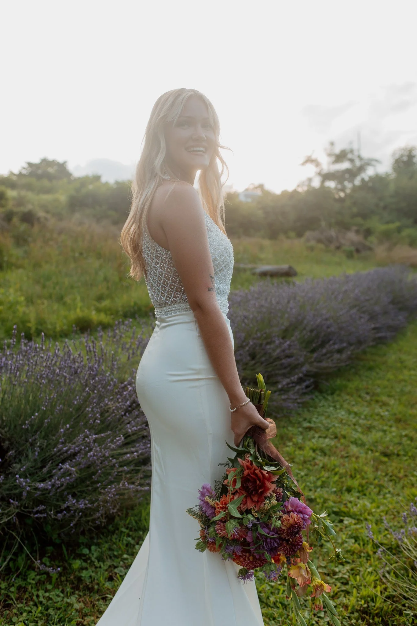 a bride holds a local flower bouquet among the lavender field while the sun sets over the flower farm