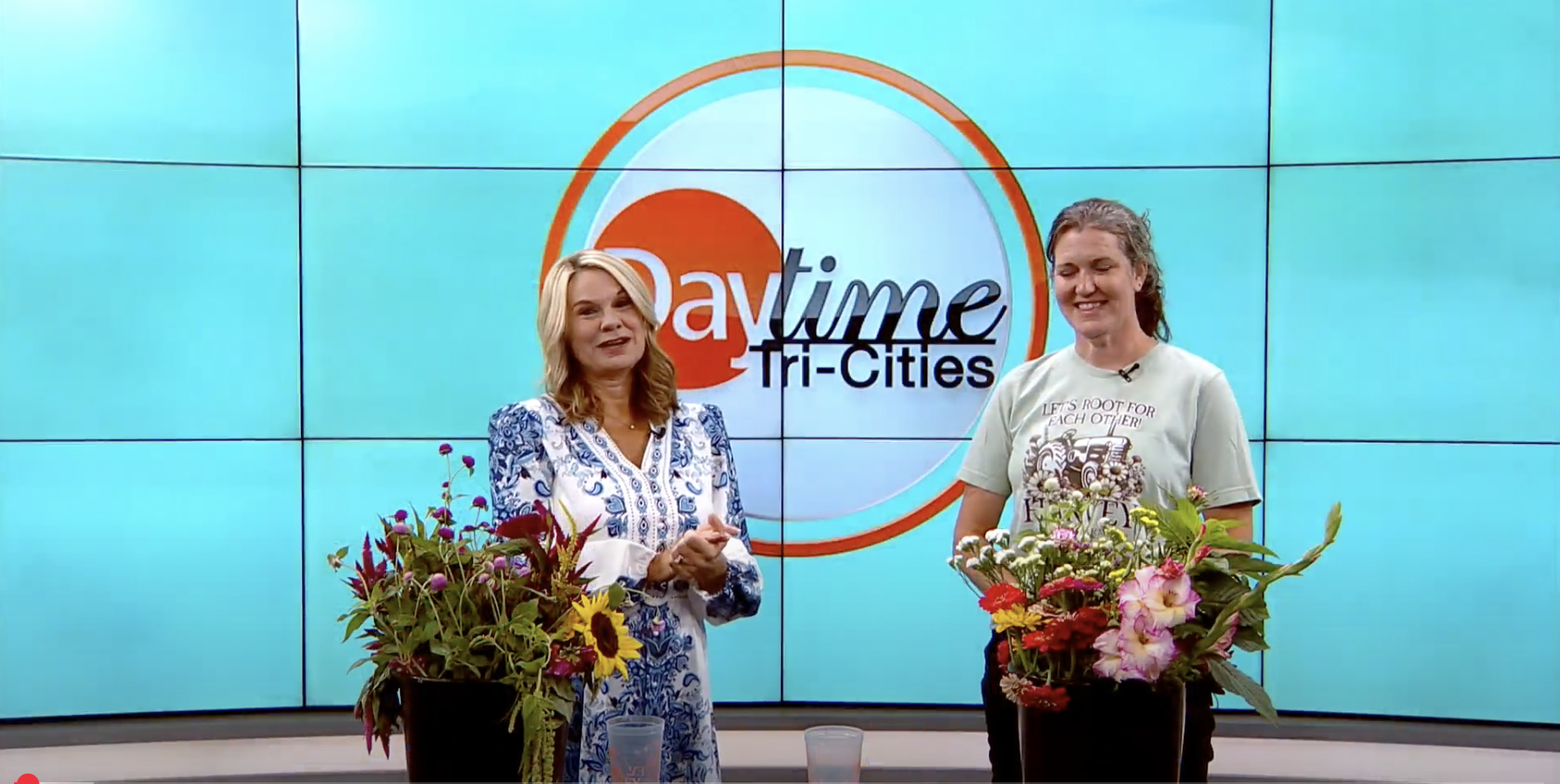 Two women standing on a TV set with a large screen behind them displaying the 'Paytme Tri-Cities' logo. Each woman is standing behind a pot of blooming flowers. The woman on the left has blonde hair and is wearing a white and blue patterned dress. Th