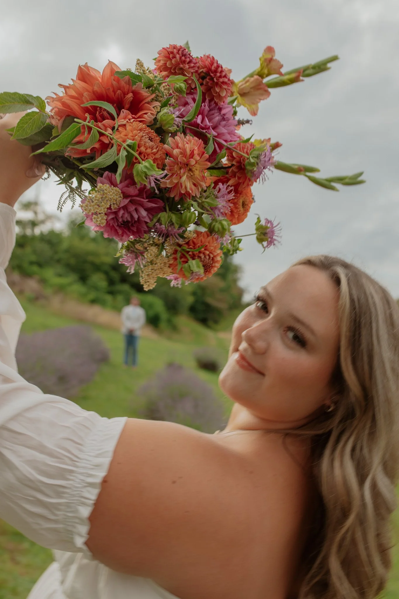 A bride holds her bouquet of fresh local flowers while her groom looks on from the lavender field