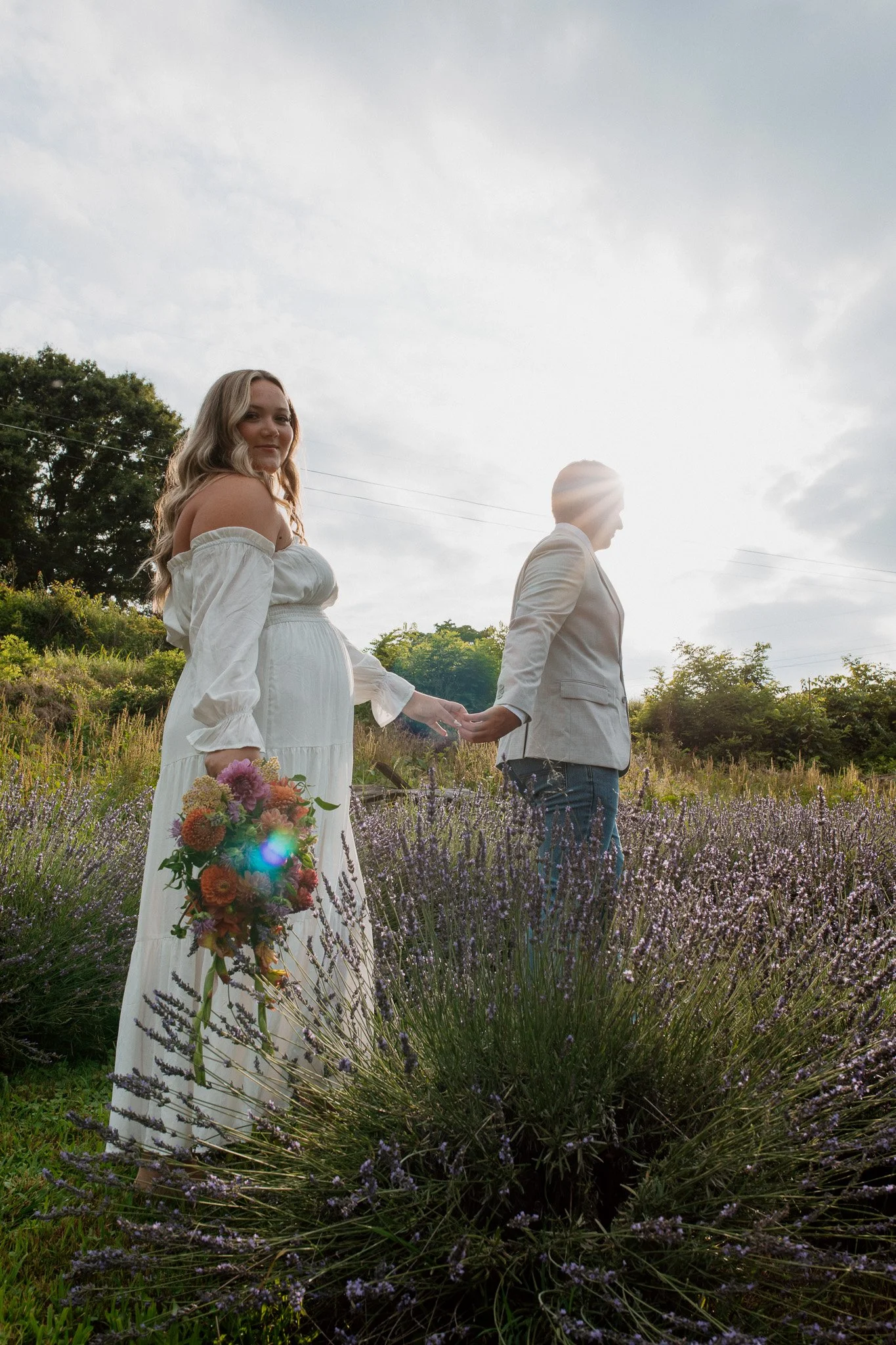 A bride looks back at the camera while holding hands with her groom.  They are in a lavender patch and she holds fresh local flowers