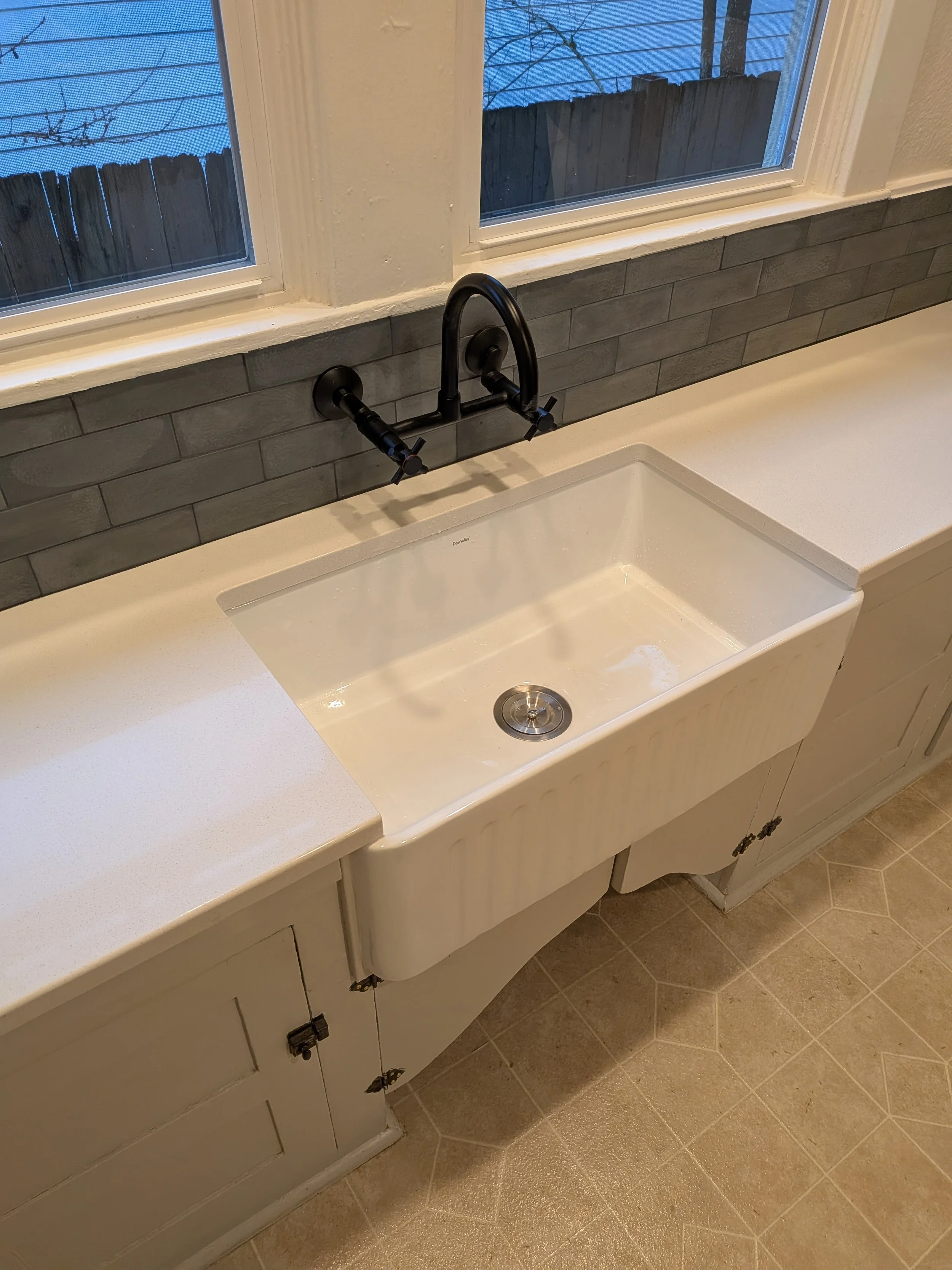 A sink with white countertop, black wall mounted faucet and grey backplash with two windows above