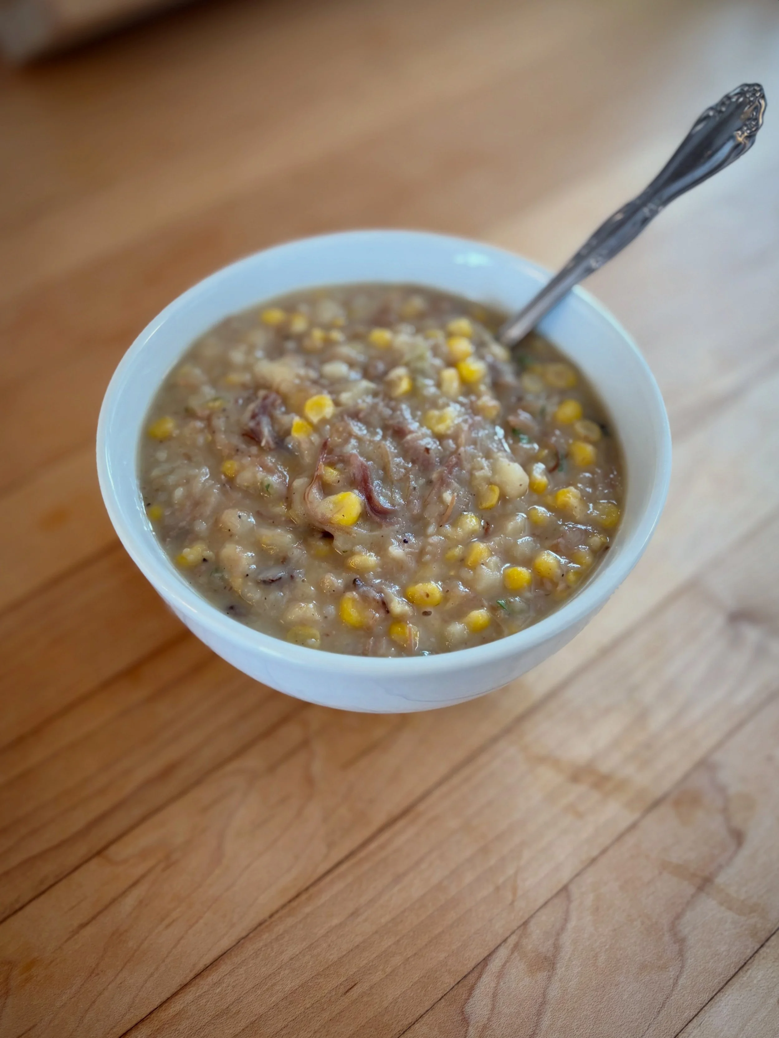 A white bowl filled with homemade-style corn and beef stew on a wooden surface, with a spoon inside the bowl.