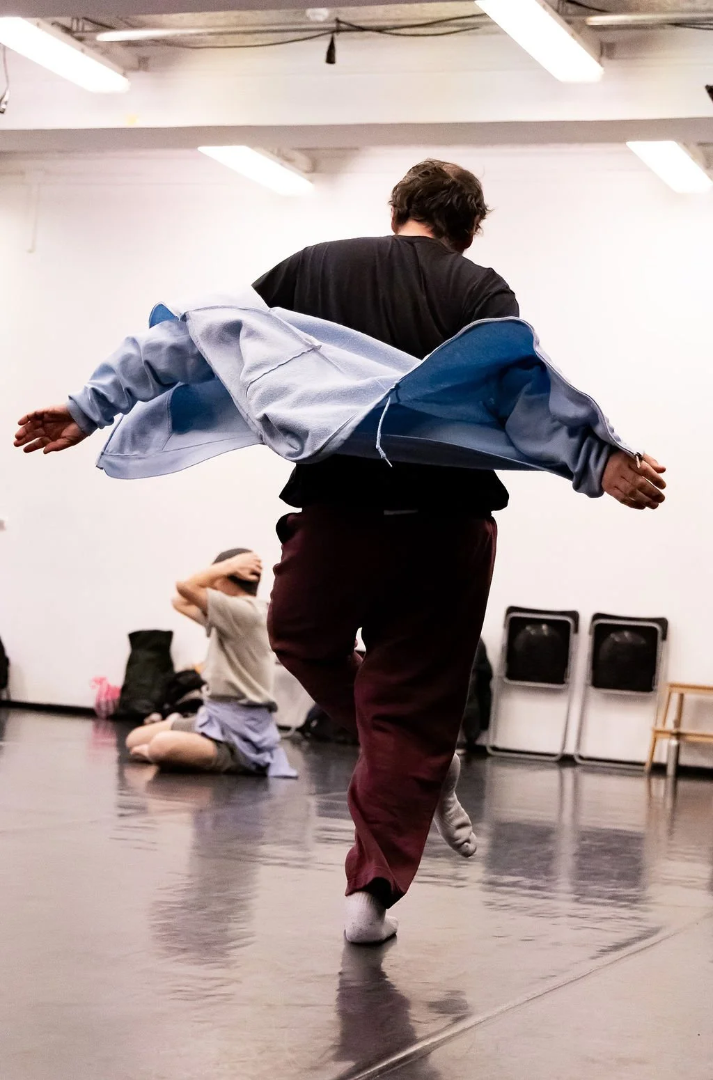 male dancer with back to camera in studio with one leg crossed over the other. Arms are splayed to the side and a blue hoodie is draped across his back. Another dancer is seated on the floor in the background, their image slightly blurred.