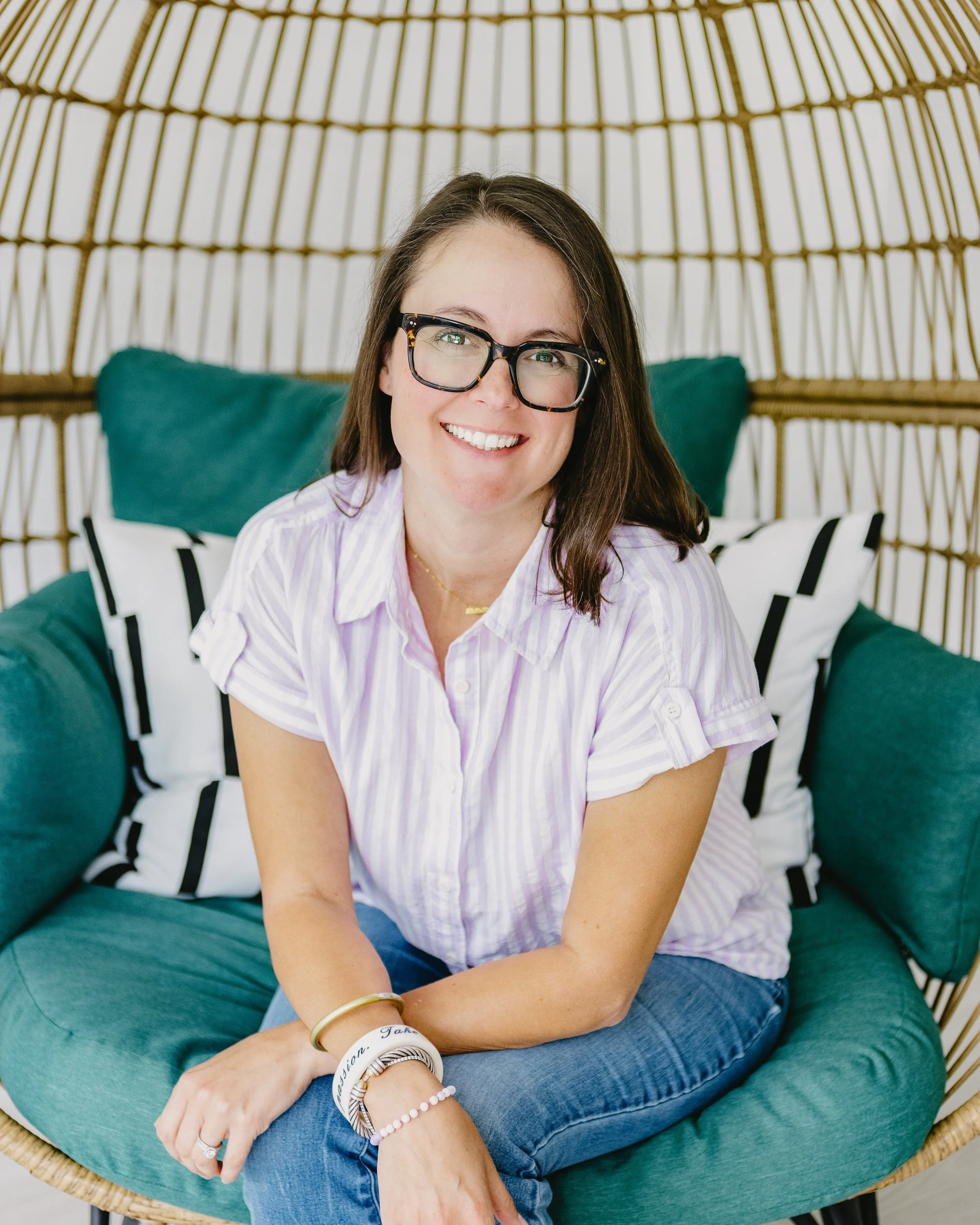 A woman with dark brown hair and glasses, smiling and sitting on a teal lounge chair with patterned pillows, in a woven rattan sunroom or patio space.