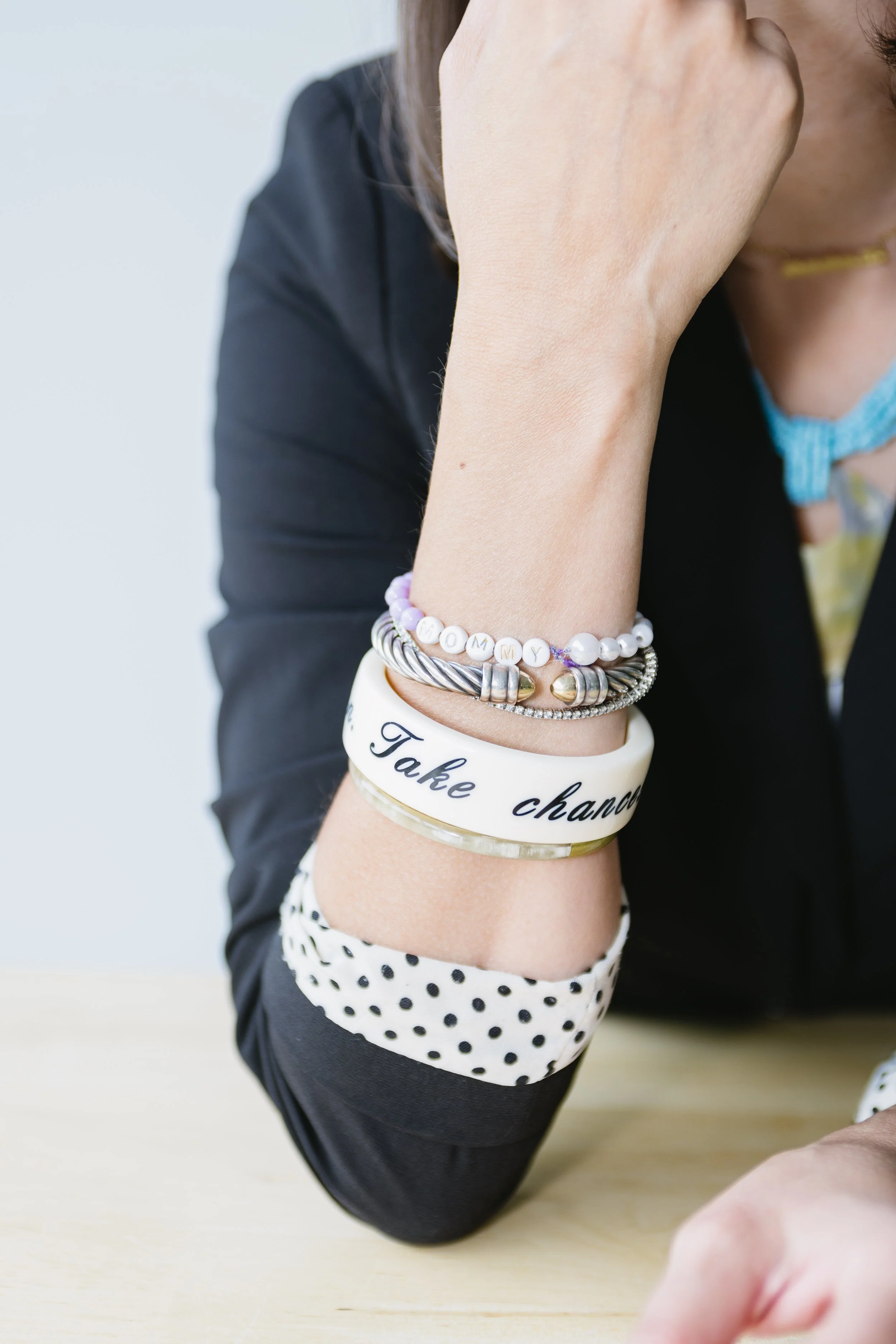 Close-up of a woman's arm resting on a table, wearing several bracelets including one with letter beads spelling 'MOMMY' and another with a quote that says 'Take chance'. She is dressed in a black blazer with polka-dot lining and a blue choker necklace.
