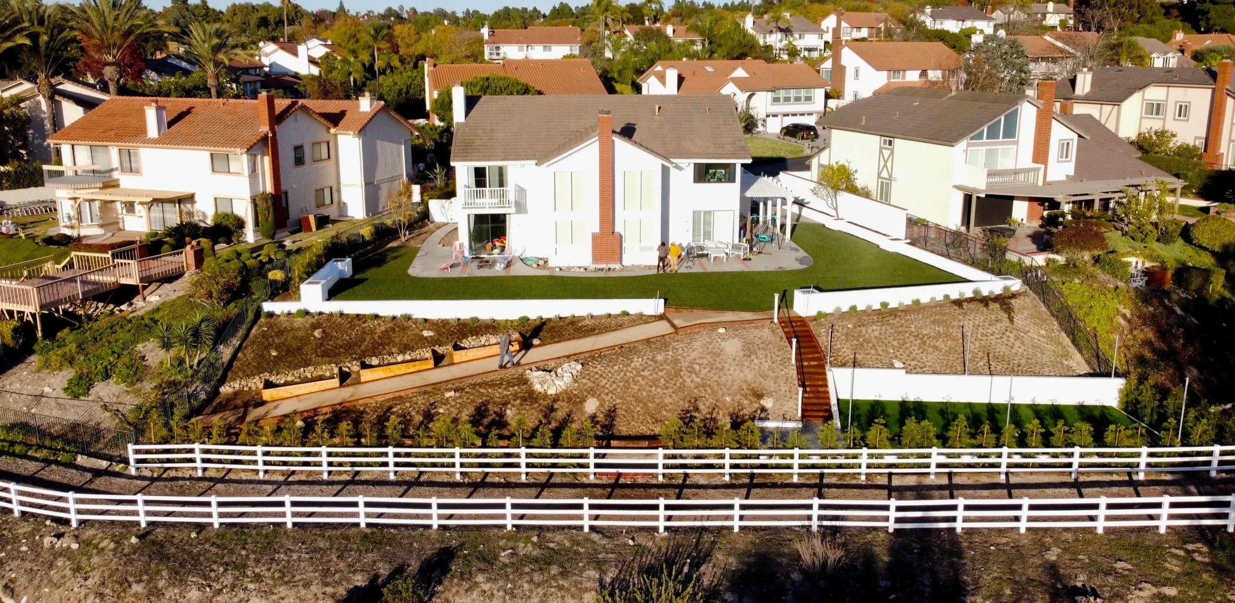 Aerial view of suburban neighborhood with houses, lawns, and landscaping. The central house has a white exterior, fenced yard, and pathways. Surrounding homes have brown roofs and are bordered by greenery and palm trees.