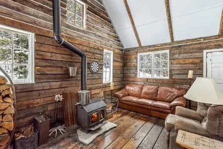 Cozy rustic living room with wooden walls, large windows, a leather sofa, an armchair, and a wood stove.