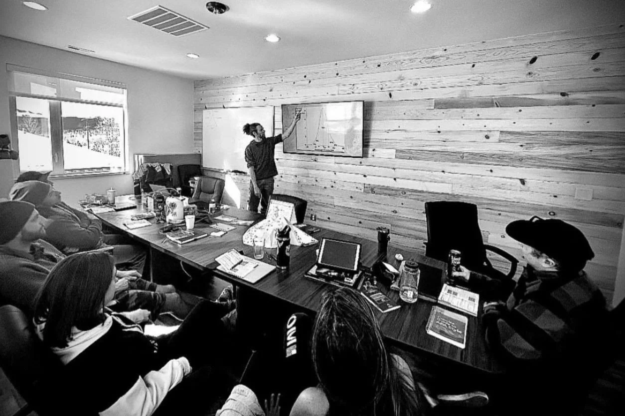 A group of people attending a meeting or class in a conference room with a wooden wall, while a person standing at the front presents information on a whiteboard and a screen.