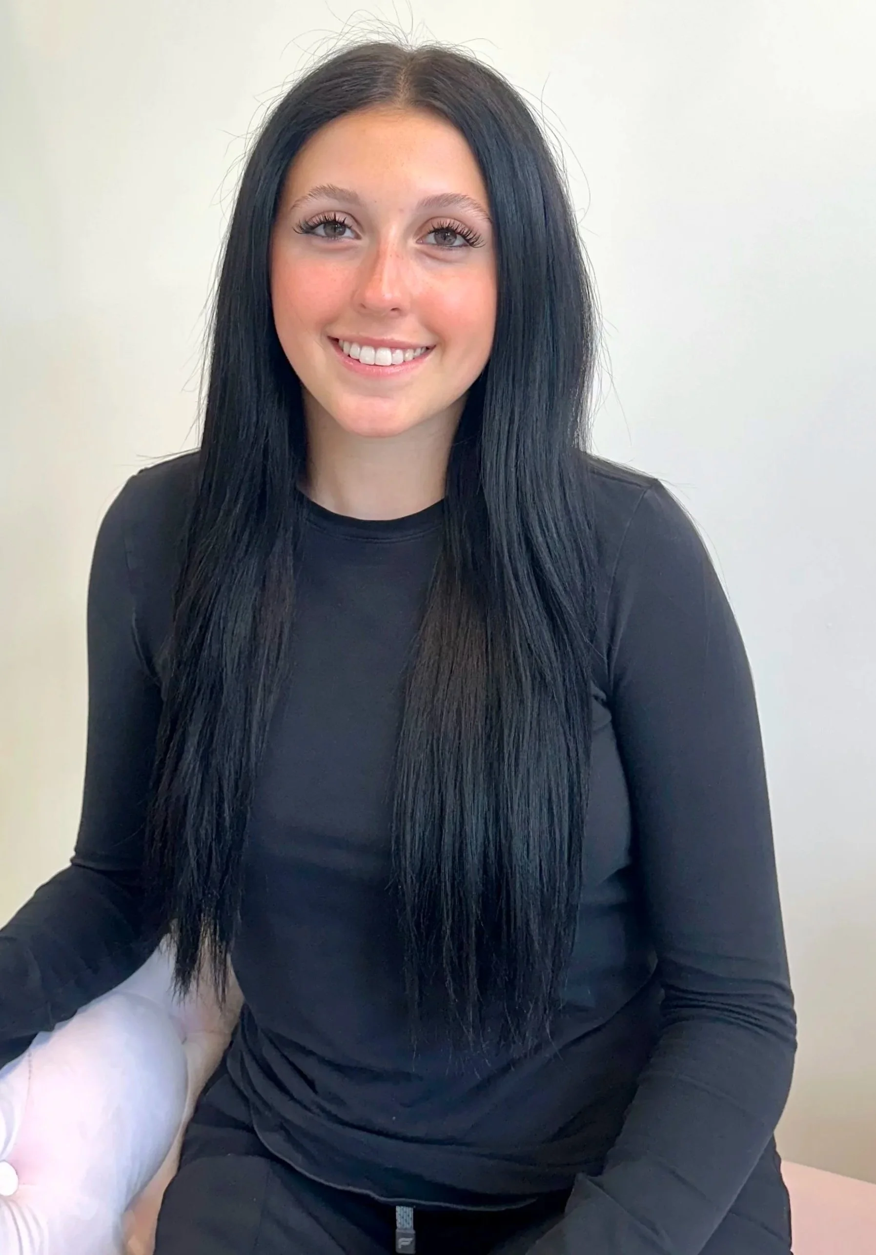 A woman with long black hair, wearing a black long sleeve shirt, sitting on a white surface against a plain white wall, smiling at the camera.