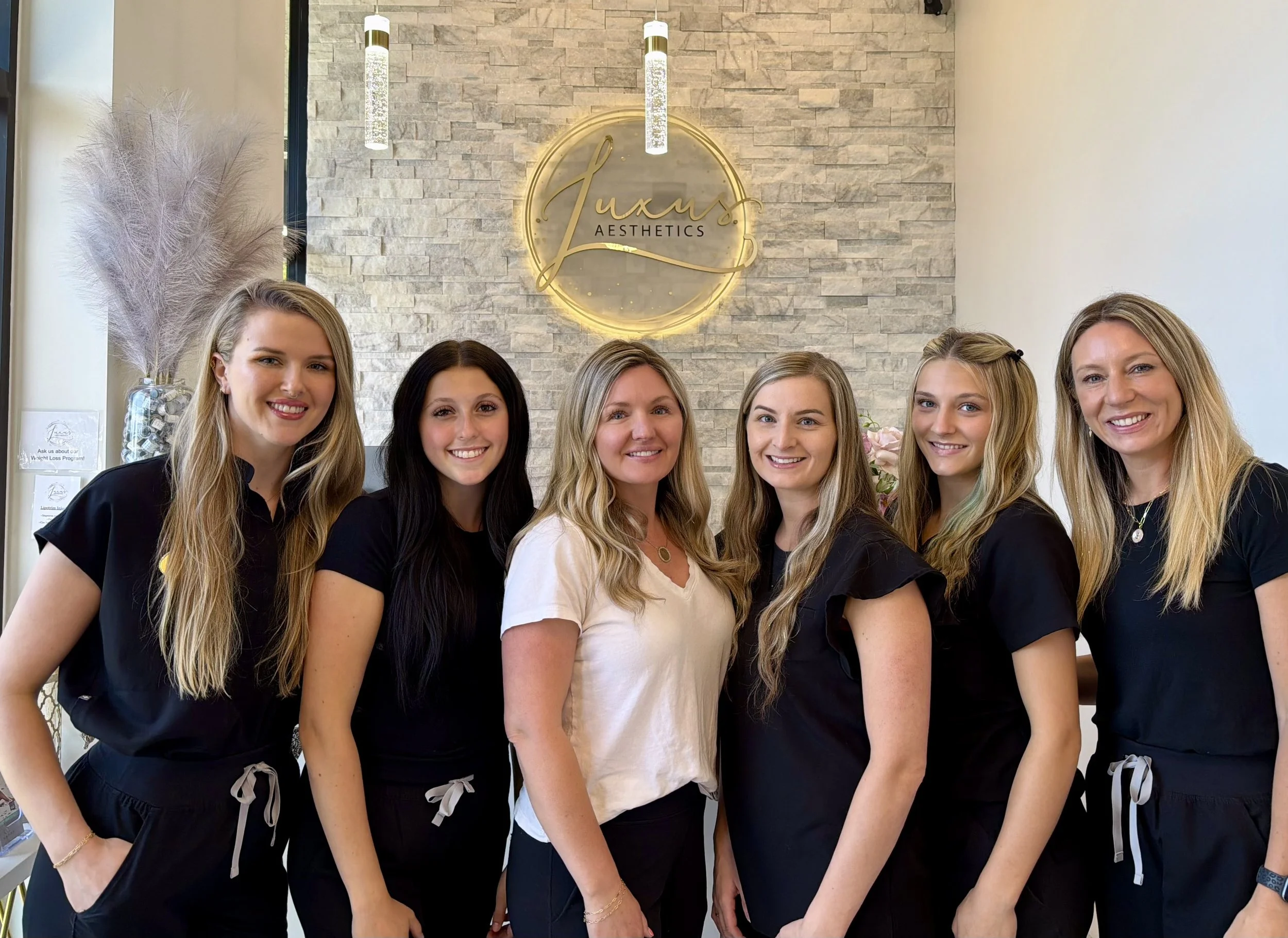 Group of six women, three with light hair and three with dark hair, standing inside a building with a stone wall and a 'Luxe Aesthetics' sign behind them. The women are smiling and dressed in black uniforms, with some wearing aprons and jewelry, in front of a decorative flower and pampas grass arrangement.