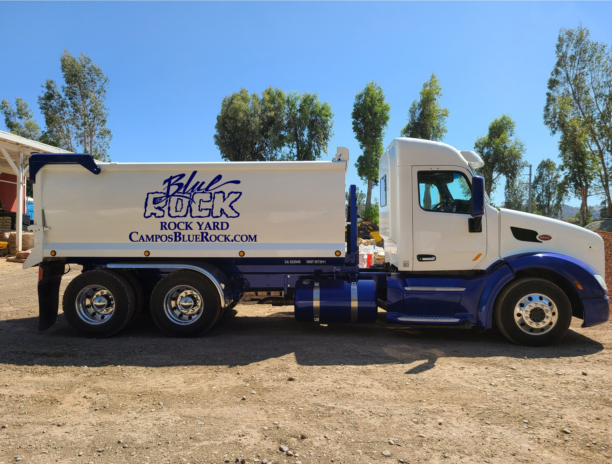 Semi-truck with white cab and blue lower section, parked on a dirt lot, with trees and a blue sky in the background. The truck has a white and blue sign on its side reading "Blue Sock Rock Yard" and a website URL.