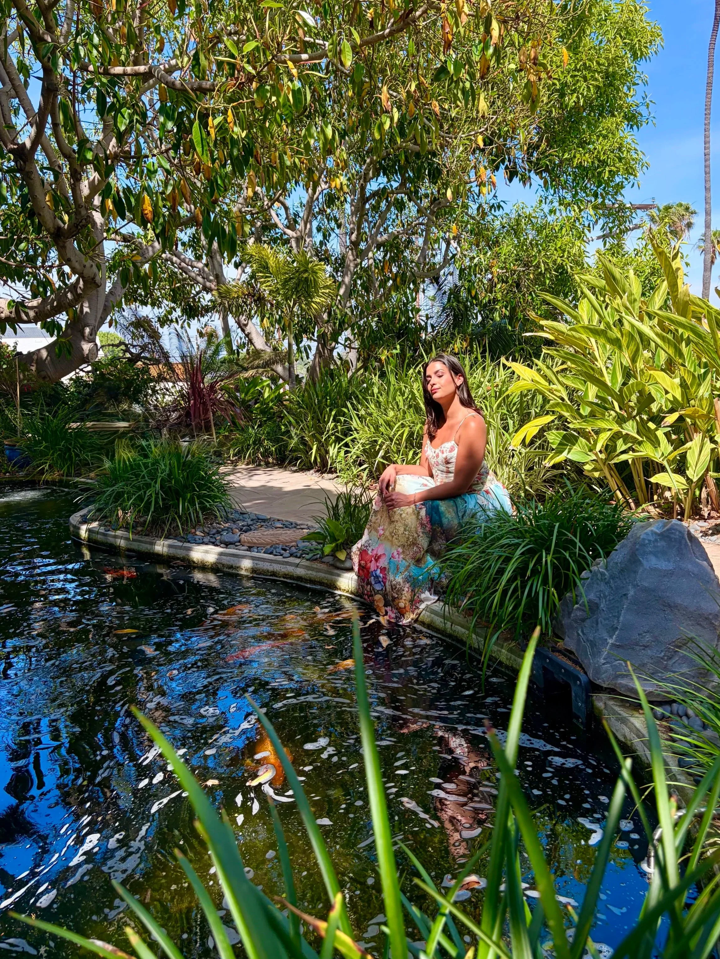 A woman sitting by a pond in a lush garden, surrounded by green plants and trees, wearing a floral dress, with blue sky in the background.