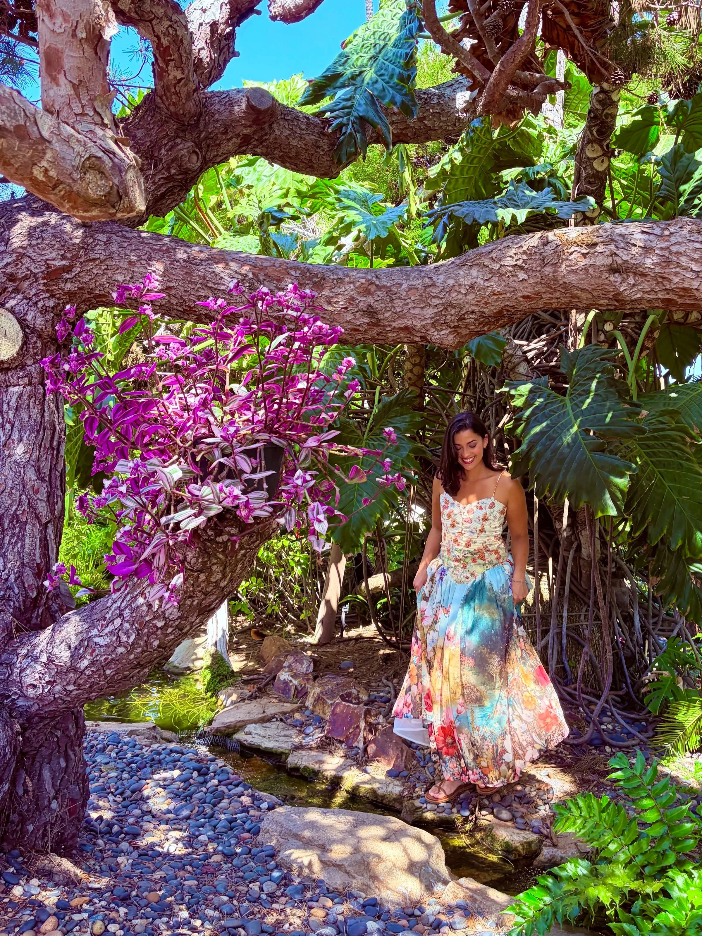Young woman in a floral dress walking on rocks near a small stream surrounded by lush green tropical plants and trees, including a large branch with pink flowers.
