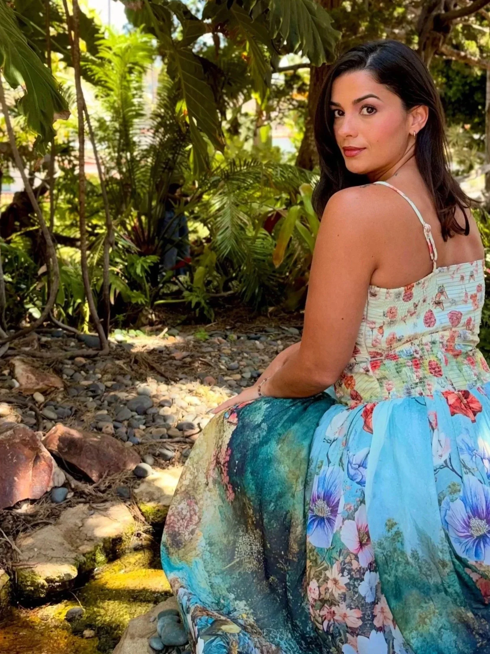 A woman sitting outdoors among lush green plants, wearing a colorful dress and looking over her shoulder at the camera.