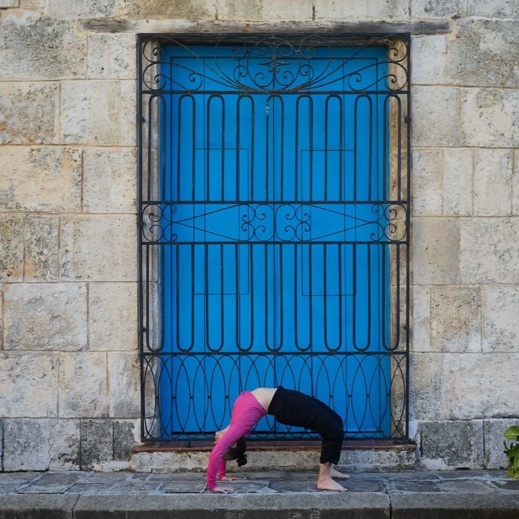 A person practicing yoga in front of a bright blue door with a black wrought iron gate, wearing a pink top and black pants, in a backbend position in Havana, Cuba. Photo by Alberto Morales.