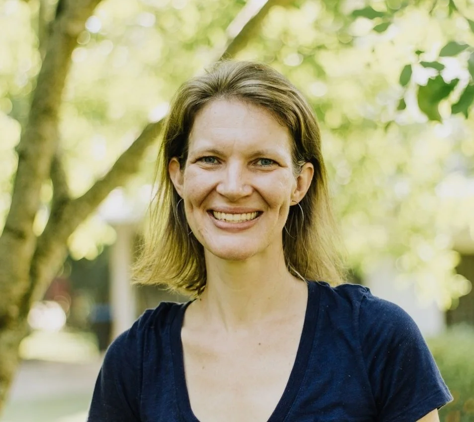A woman with shoulder-length brown hair smiling outdoors with trees and greenery in the background.