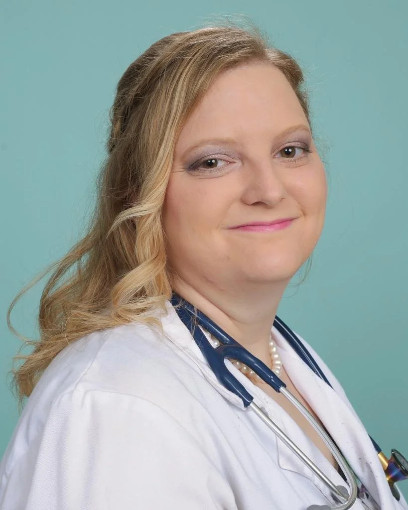 A smiling female healthcare professional with blonde hair, wearing a white coat and a stethoscope around her neck, posed against a light teal background.