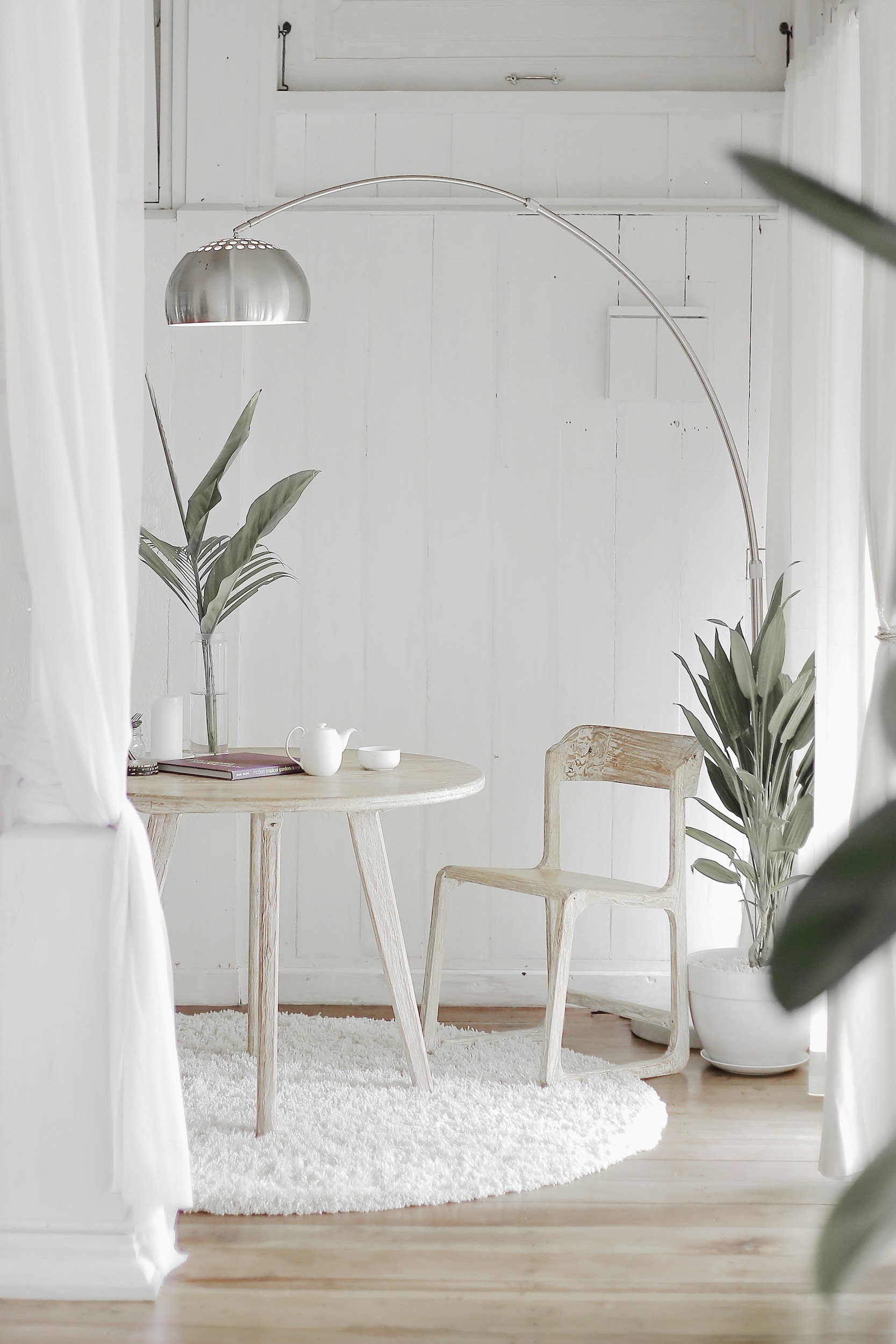 A bright, minimalist room with white walls and wooden floors, featuring a rustic wooden chair, a white rug, and potted plants. A large silver arc floor lamp hangs over a round wooden table with a white teapot, cup, and a few books.