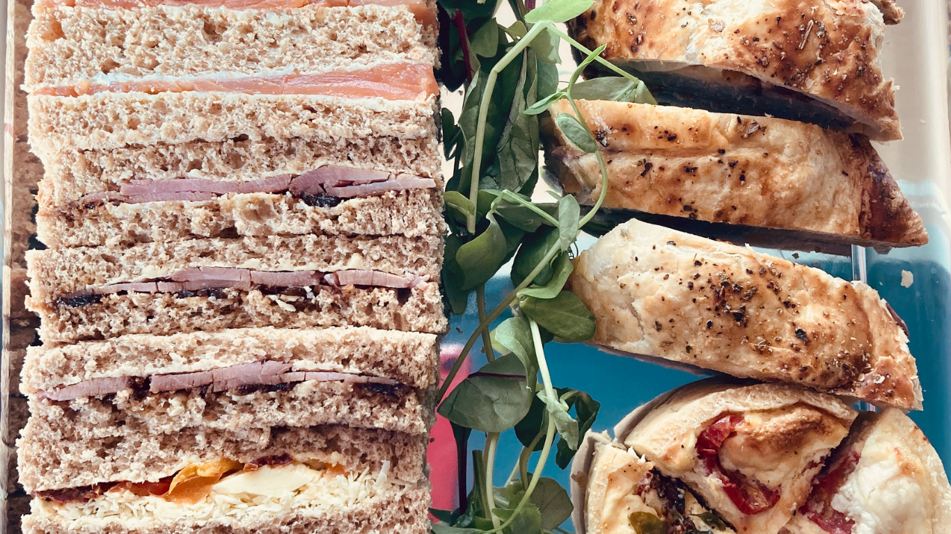 A tray with sliced sandwiches, grilled chicken pieces, and a pizza slice with toppings including cheese, tomatoes, and peppers, garnished with leafy greens.